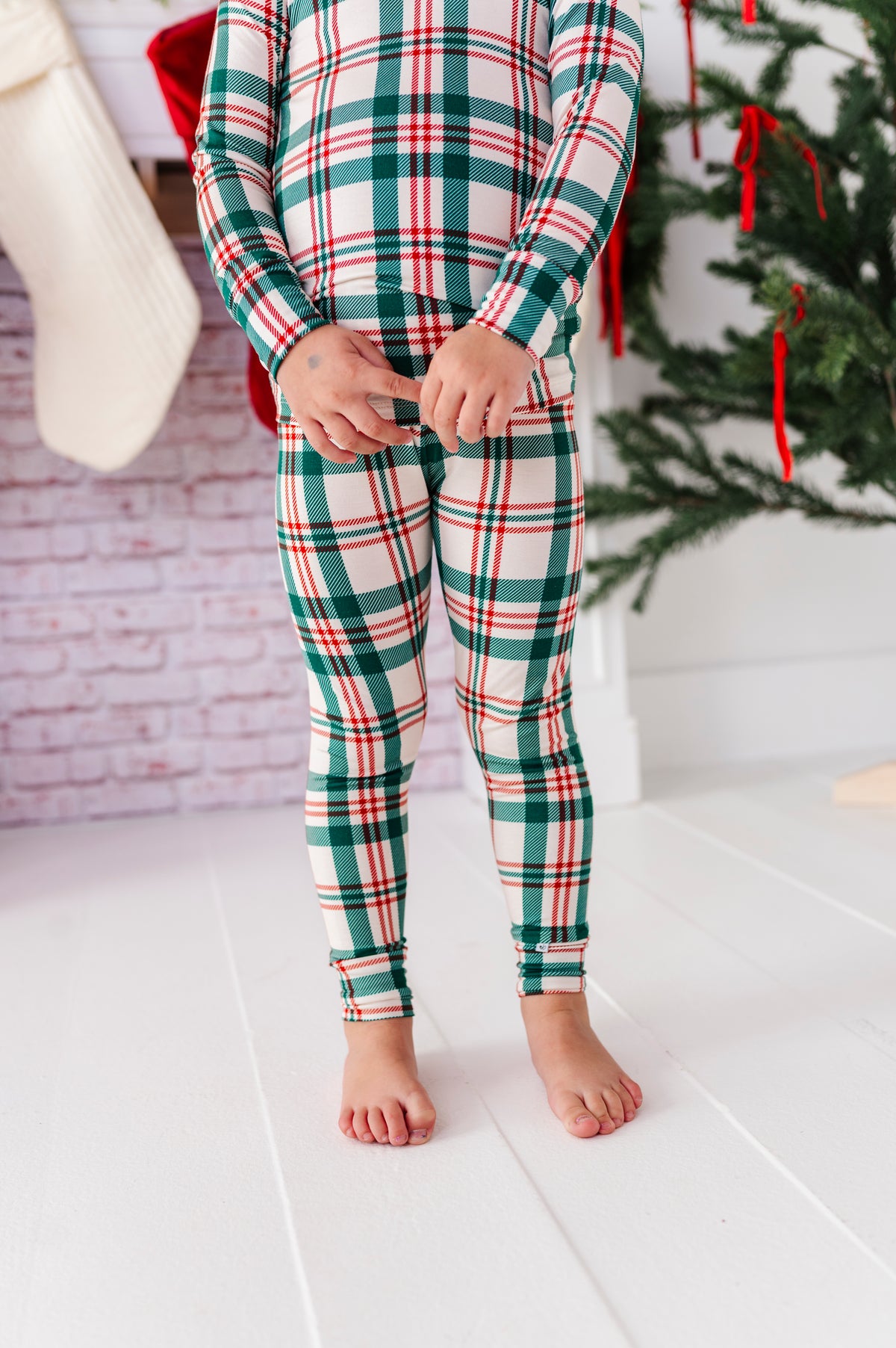 A close-up shot of the Classic Christmas plaid pajama set with a big red bow in her hair, standing in front of a decorated fireplace and a Christmas tree