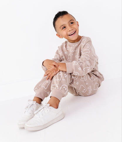 A young boy smiling and laughing while wearing a matching beige pajama set with white cartoon patterns, sitting on a white floor against a white wall.