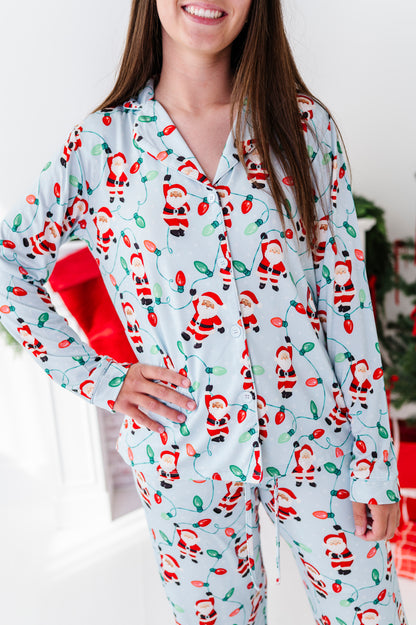 A woman wearing a light blue long-sleeve bamboo pajama set with a festive Santa Claus and Christmas light print, holding a red gift box in a bright indoor setting.