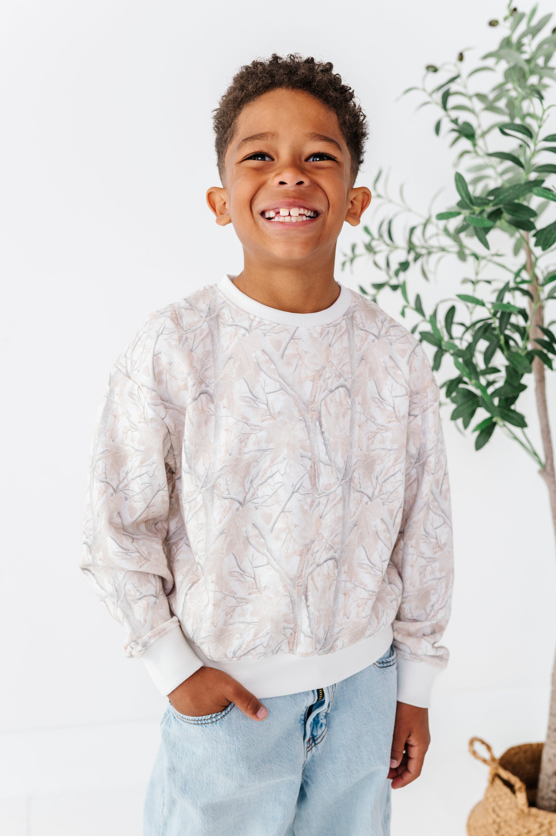 Child wearing a light-colored camo patterned sweater and jeans standing indoors with a plant in the background.