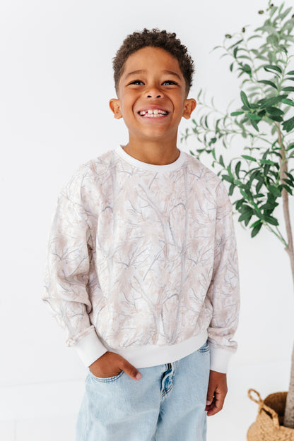 Child wearing a light-colored camo patterned sweater and jeans standing indoors with a plant in the background.