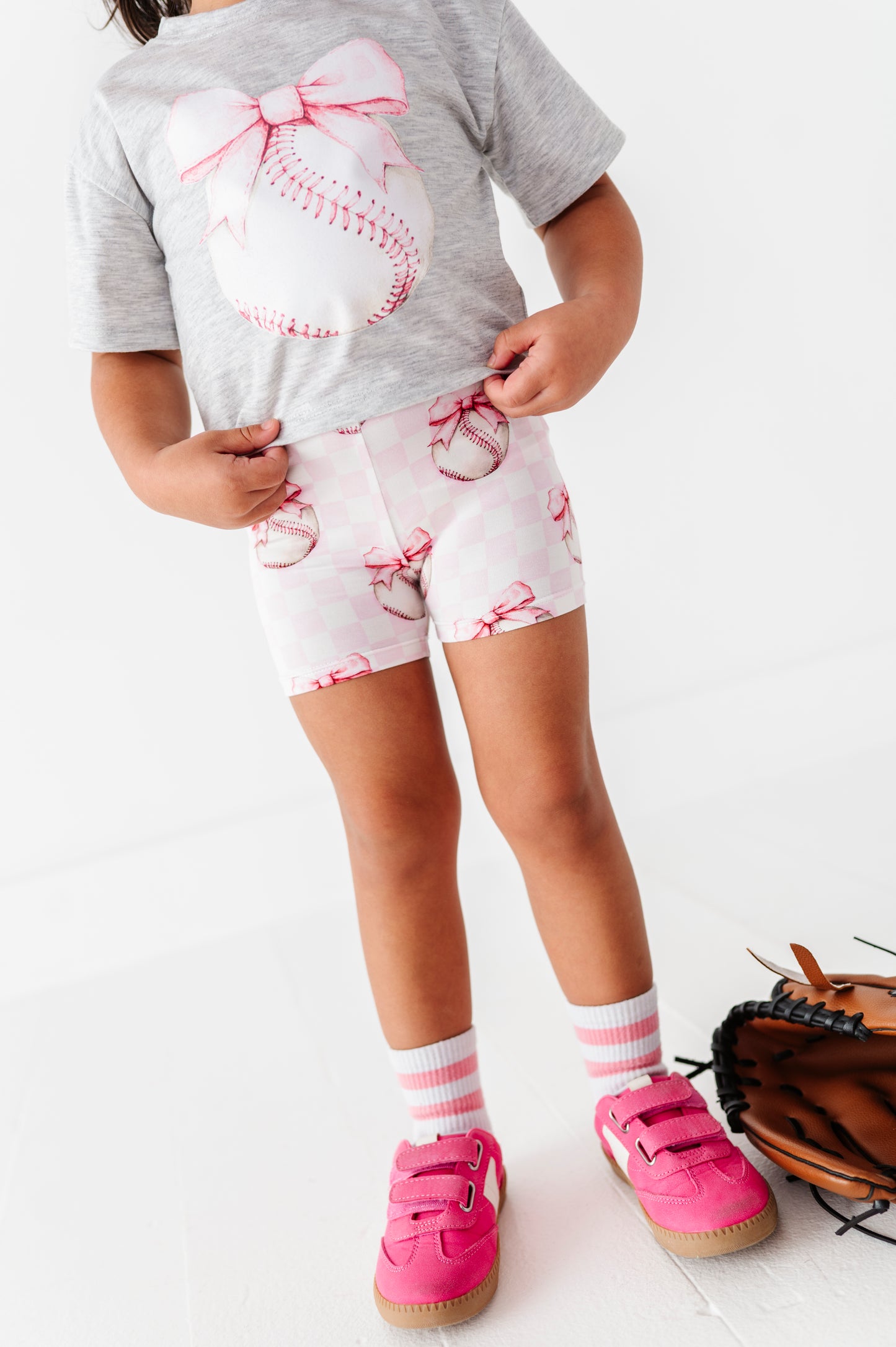 Child wearing a gray t-shirt with a baseball design, pink bamboo shorts, pink shoes, and striped socks on a white background.
