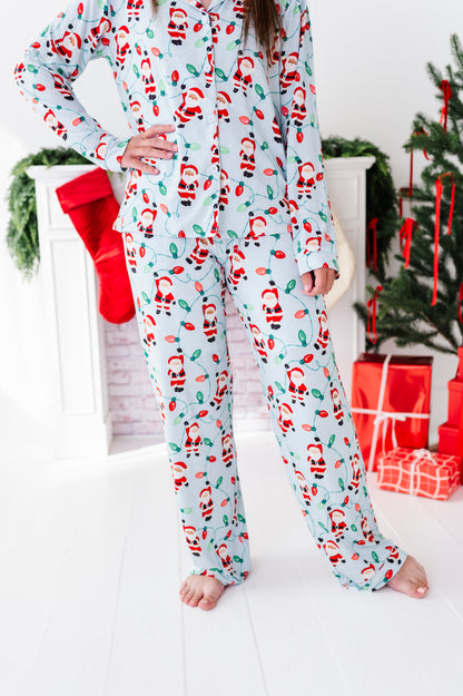 A woman wearing a light blue long-sleeve bamboo pajama set with a red and green Santa Claus print, standing barefoot in a holiday-themed room with a Christmas tree, stockings, and wrapped gifts in the background.