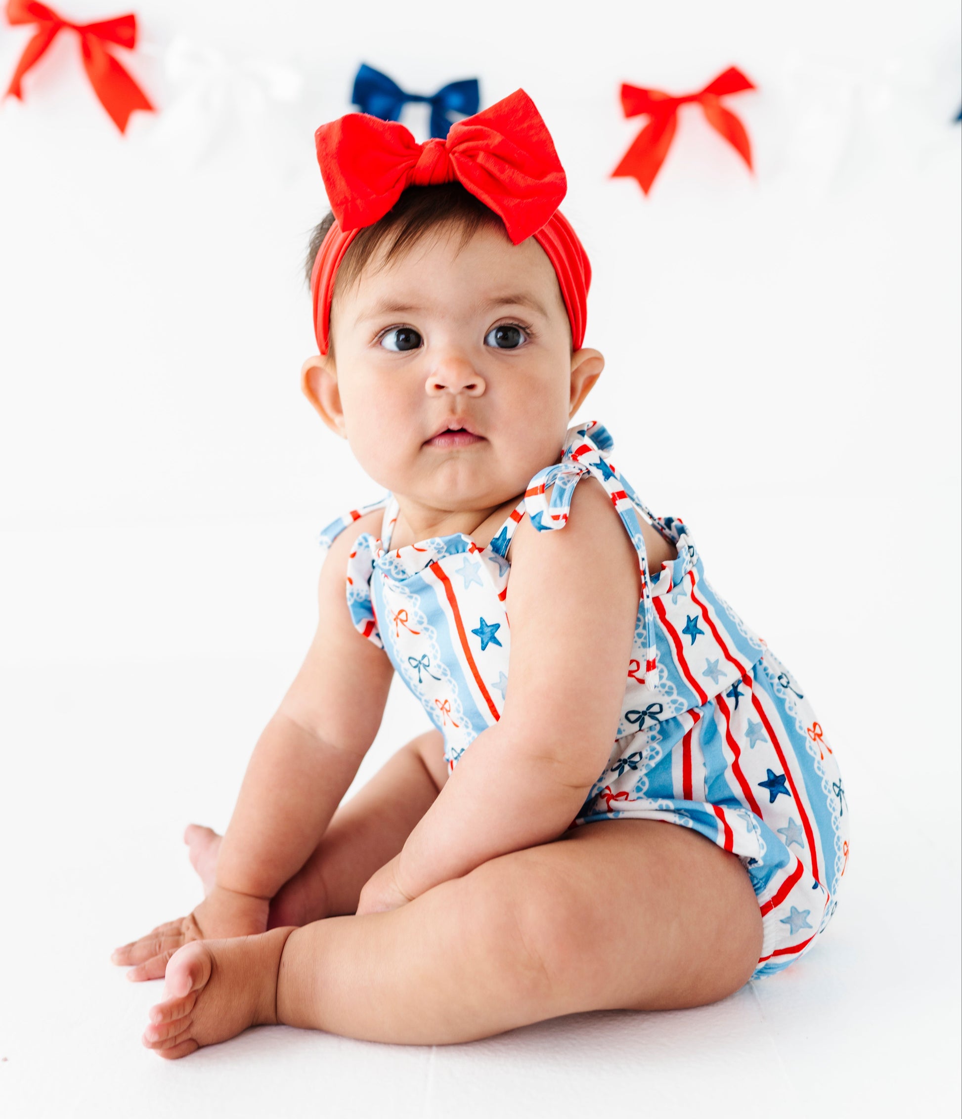Baby wearing a red bow headband and a 4th of July themed bubble romper with a white background