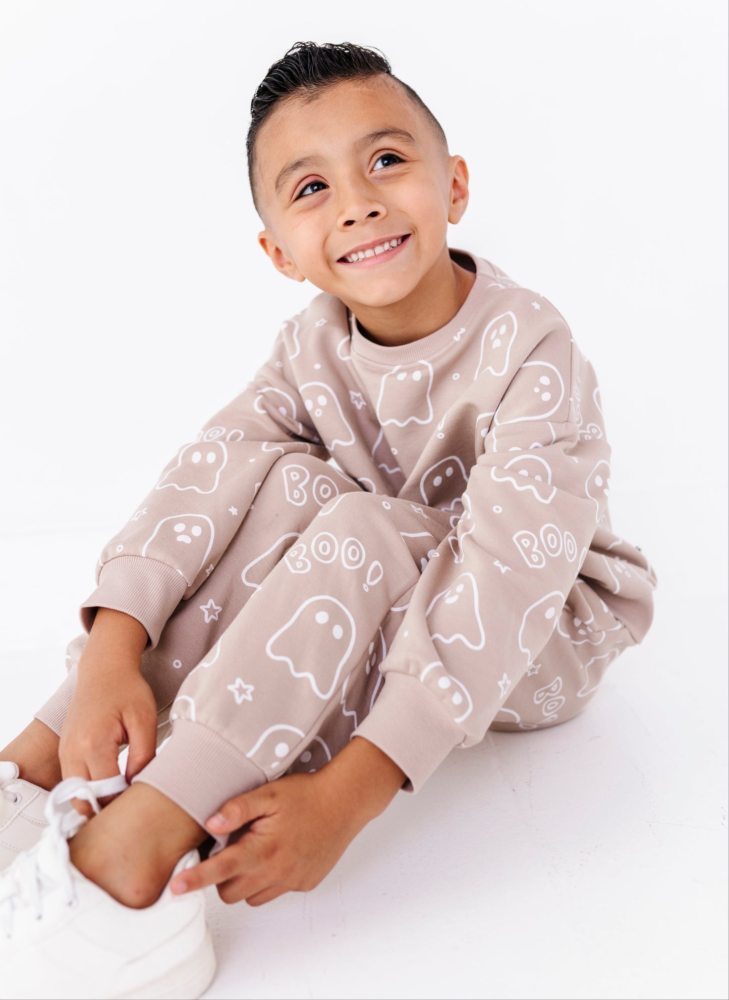 A young boy sitting on a white floor wearing a matching beige pajama set with white ghost and star patterns, paired with white sneakers.