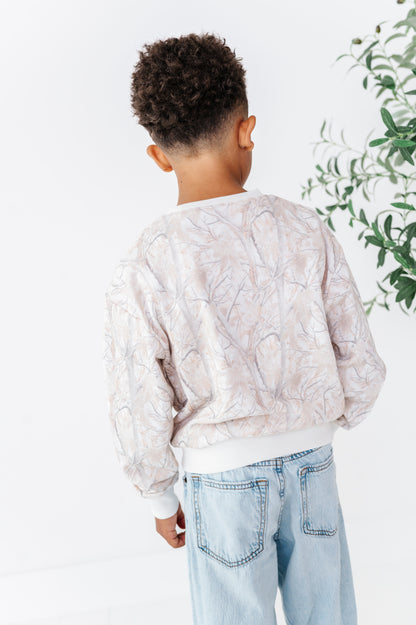 Person wearing a camo patterned sweatshirt  and light blue jeans standing against a white wall with greenery.