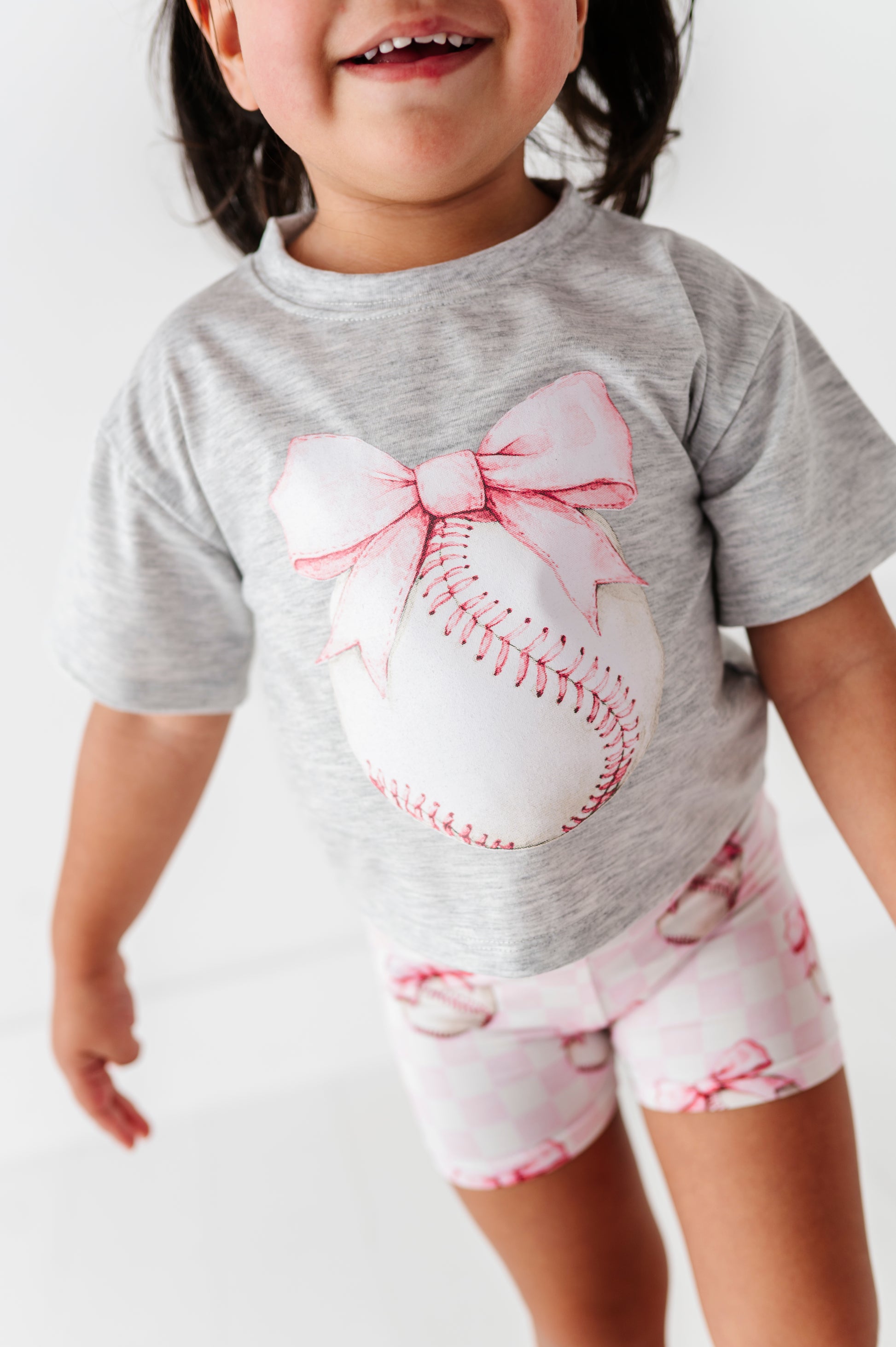 Child wearing a gray t-shirt with a baseball and bow design and pink checkered bamboo shorts 