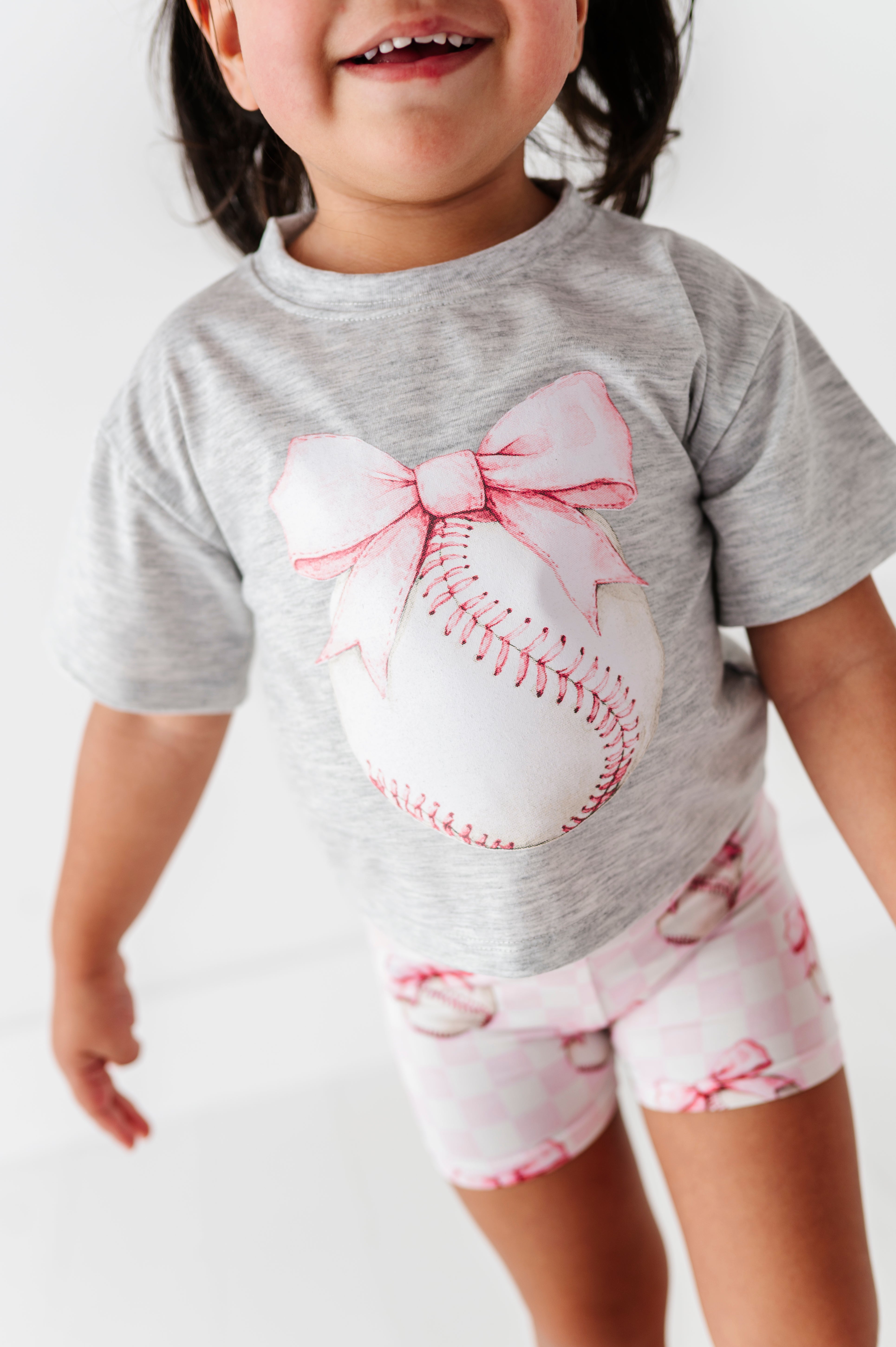 Child wearing a gray t-shirt with a baseball and bow design and pink checkered bamboo shorts 