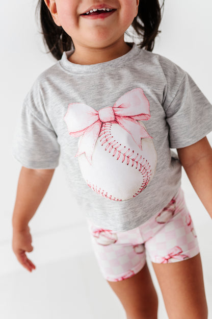 Child wearing a gray t-shirt with a baseball and bow design and pink checkered bamboo shorts 