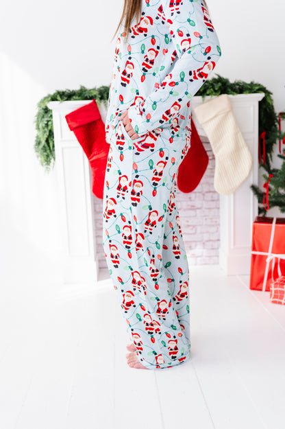 A person wearing a light blue Christmas bamboo pajama set with a Santa Claus print, standing in front of a white fireplace decorated with stockings and holiday greenery.