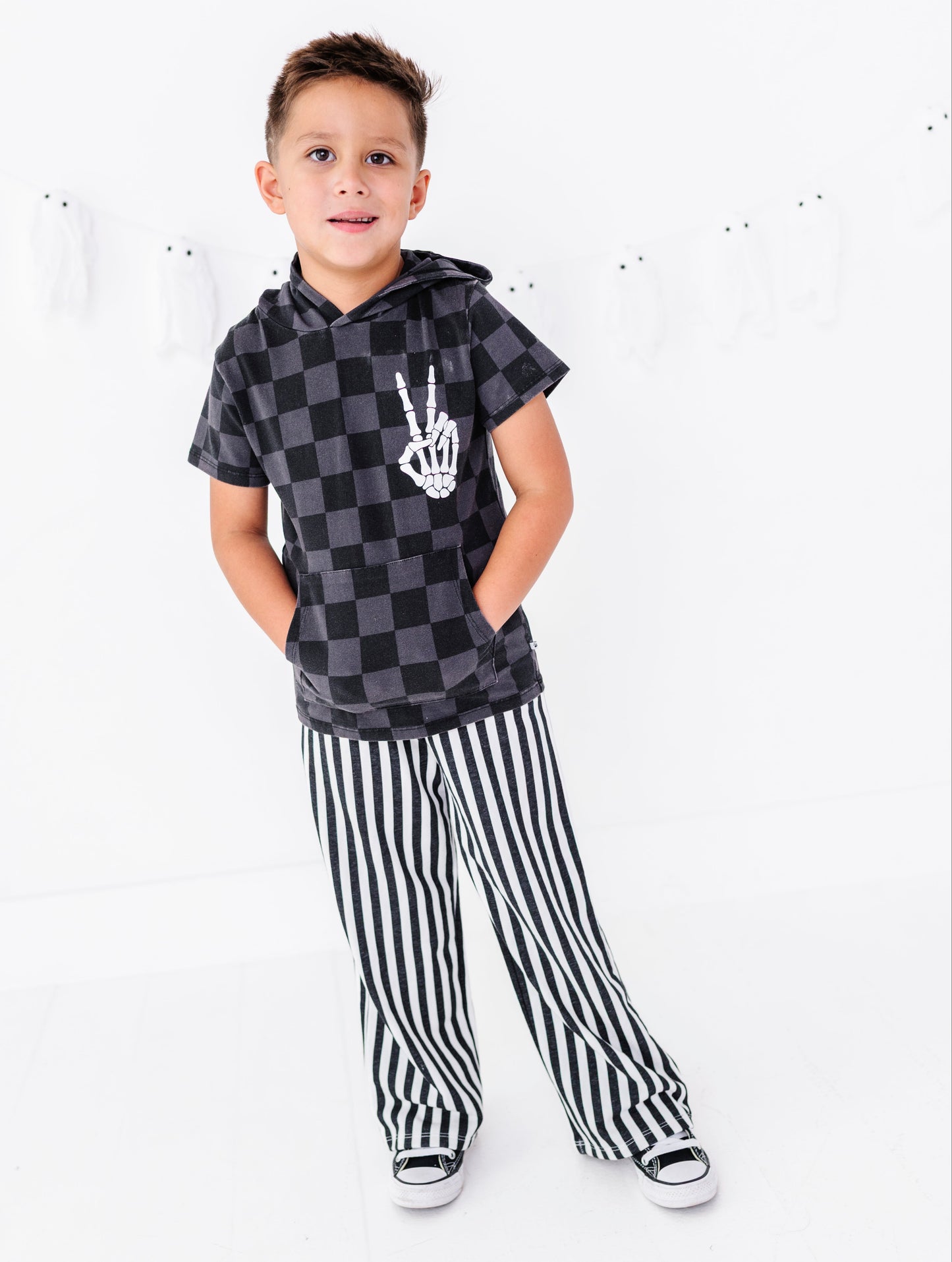 A young boy modeling a black and gray checkered hoodie with a white skeleton hand graphic and black-and-white vertically striped pants, standing against a white studio background with ghost decorations.