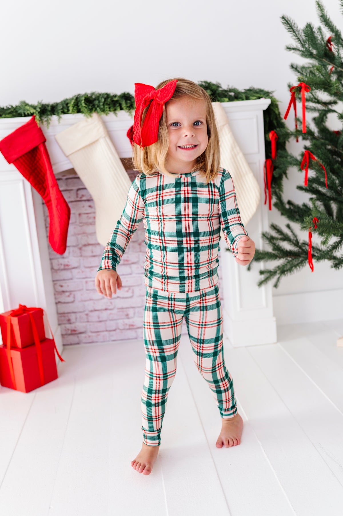 A little girl wearing a Classic Christmas plaid pajama set with a big red bow in her hair, standing in front of a decorated fireplace and a Christmas tree
