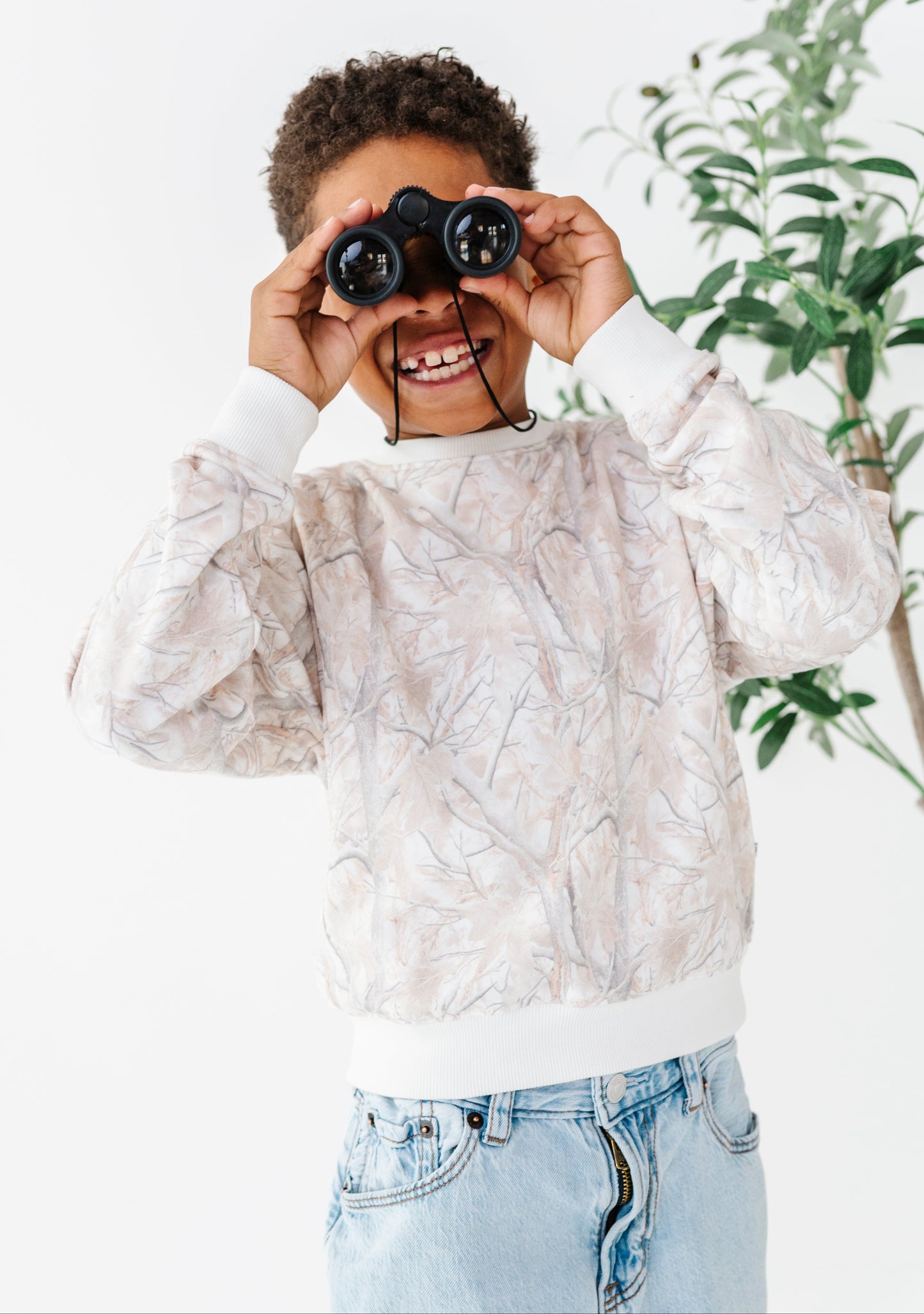 Child holding binoculars up to their eyes with a white background and plant in the corner