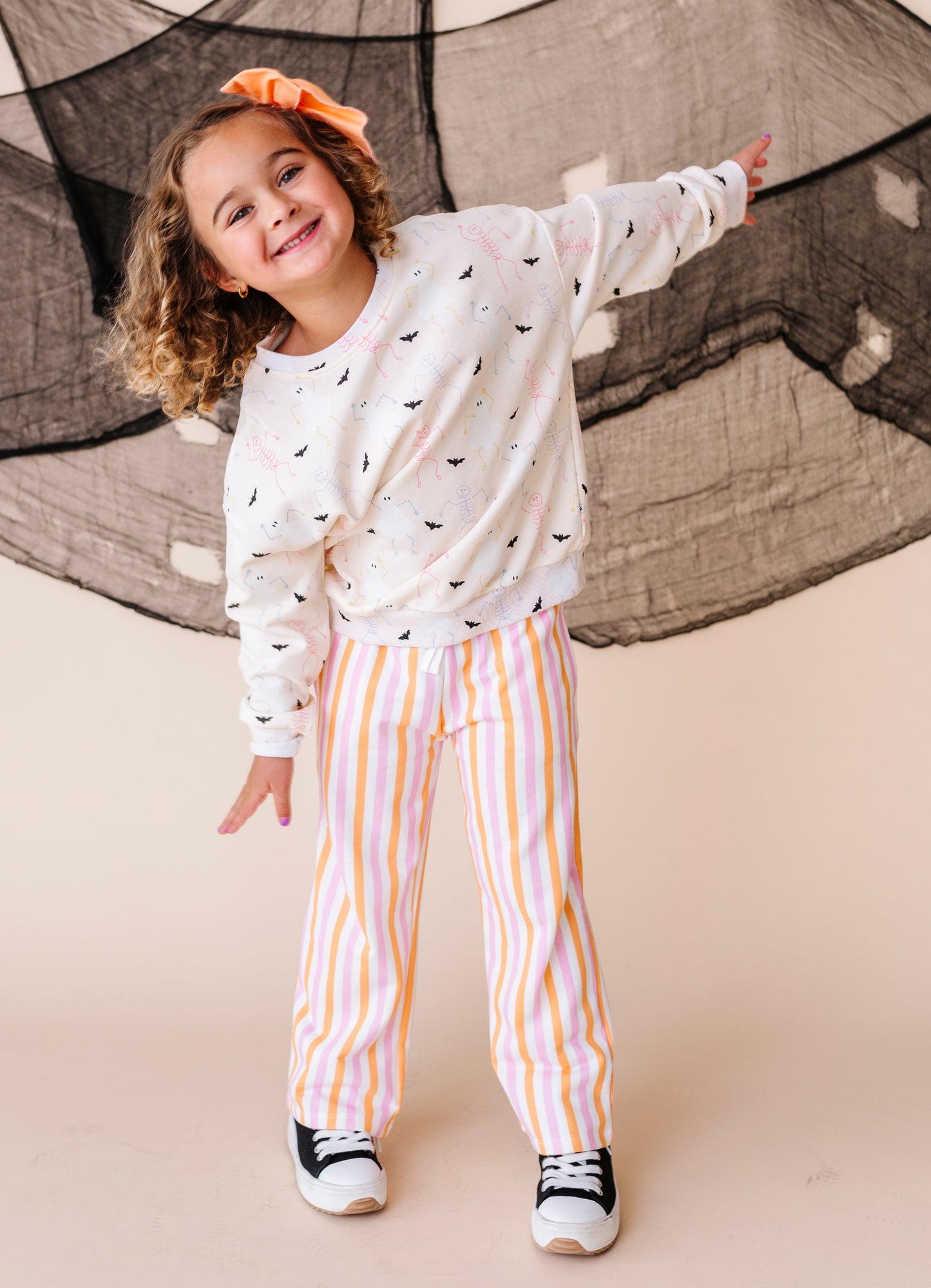 Child wearing a white long-sleeve shirt with black patterns and striped pants, standing against a neutral background.
