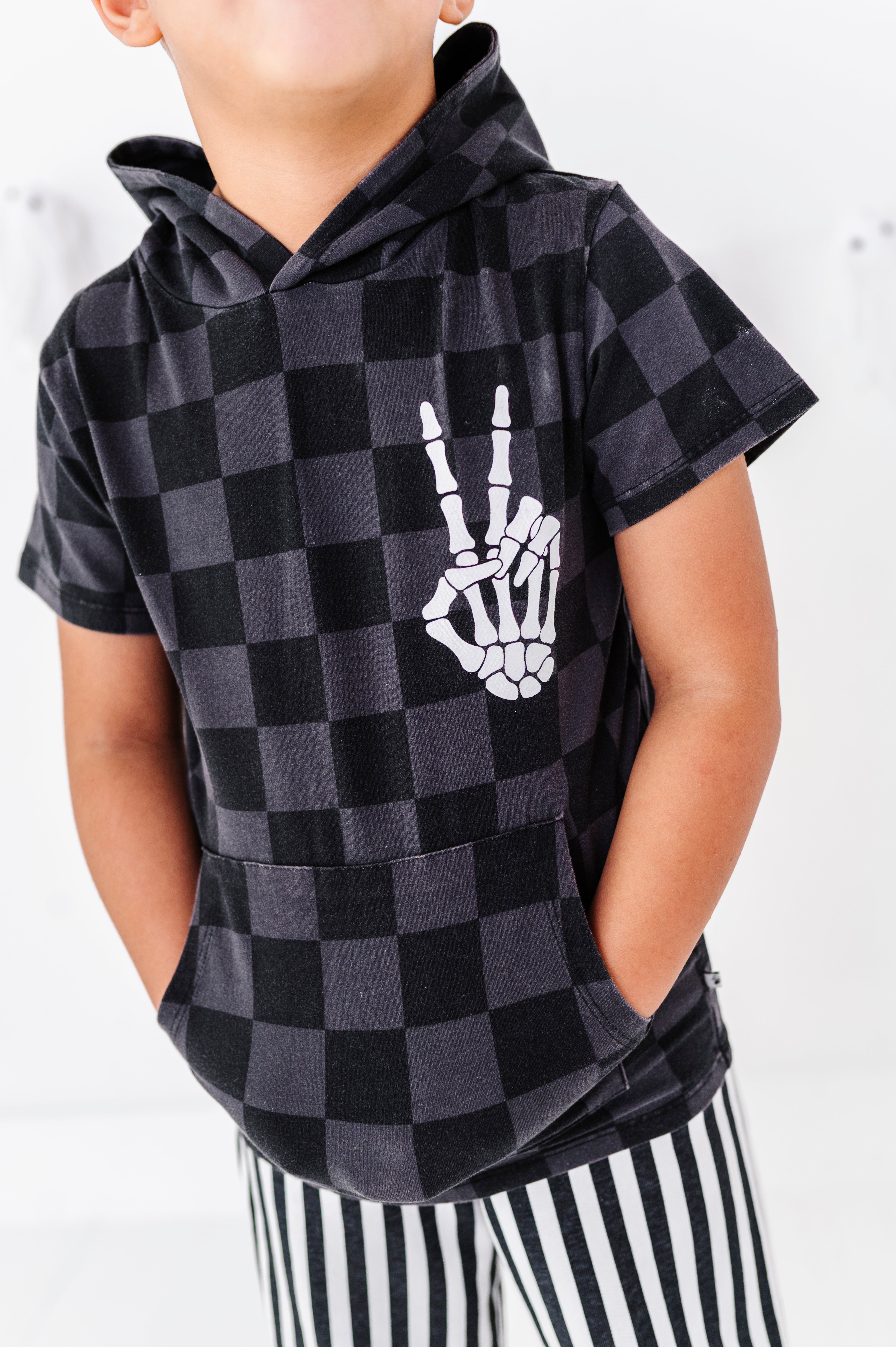 A child wearing a black and dark gray checkered hoodie with a white skeleton hand graphic on the chest, paired with black and white striped pants.