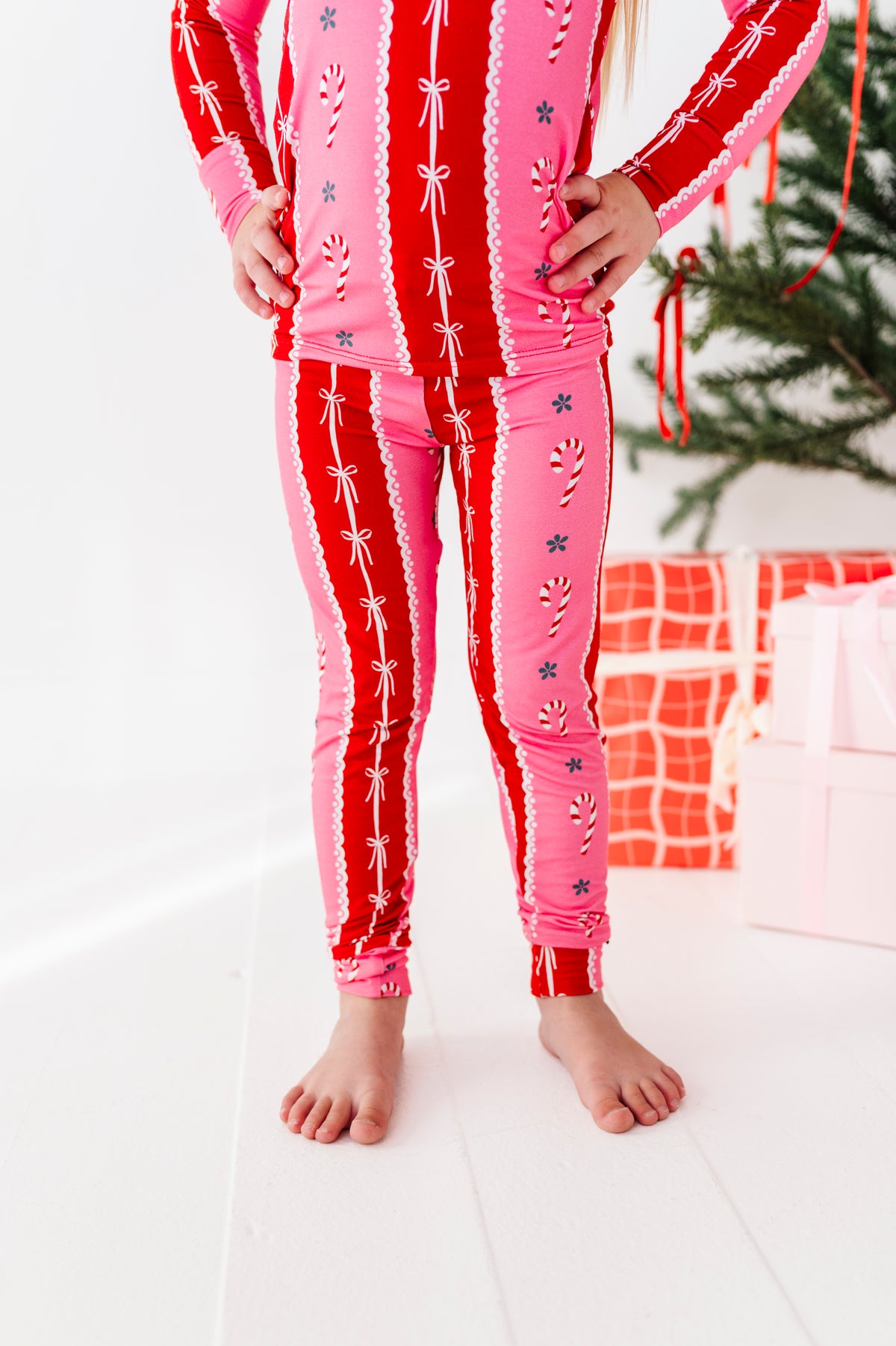 A child wearing a matching pink and red holiday bamboo pajama set with candy cane and bow patterns, standing barefoot on a white floor near wrapped gifts and a Christmas tree.