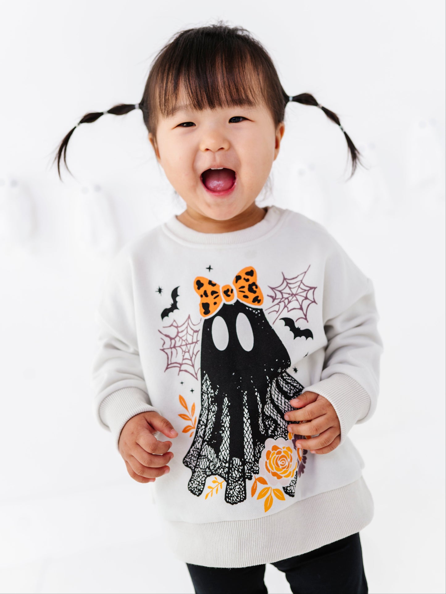 A young child with dark pigtails wearing a white long-sleeve sweatshirt with a black ghost graphic decorated with an orange bow, spiderwebs, bats, and orange leaves, set against a bright white background.