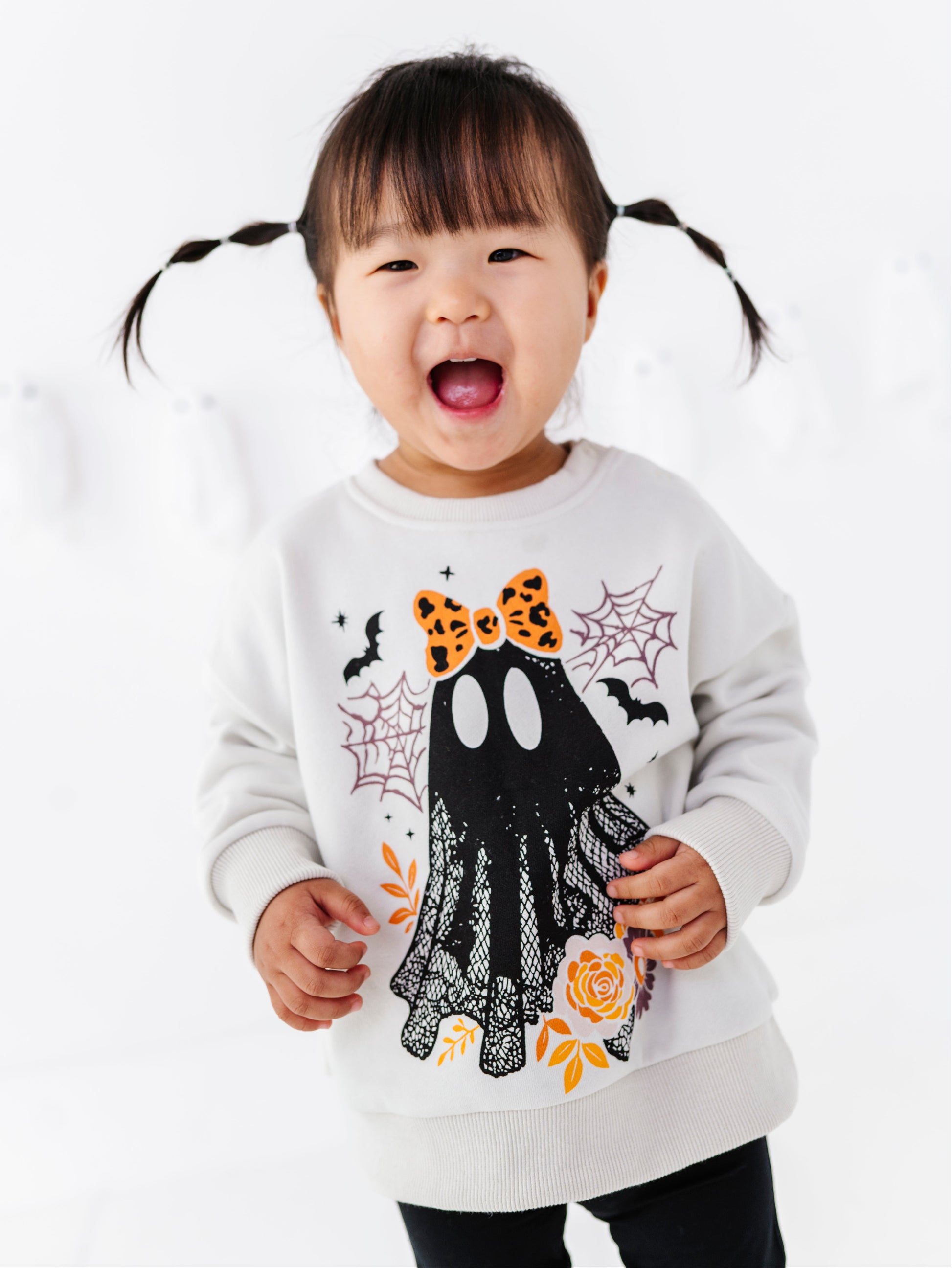 A young child with dark pigtails wearing a white long-sleeve sweatshirt with a black ghost graphic decorated with an orange bow, spiderwebs, bats, and orange leaves, set against a bright white background.