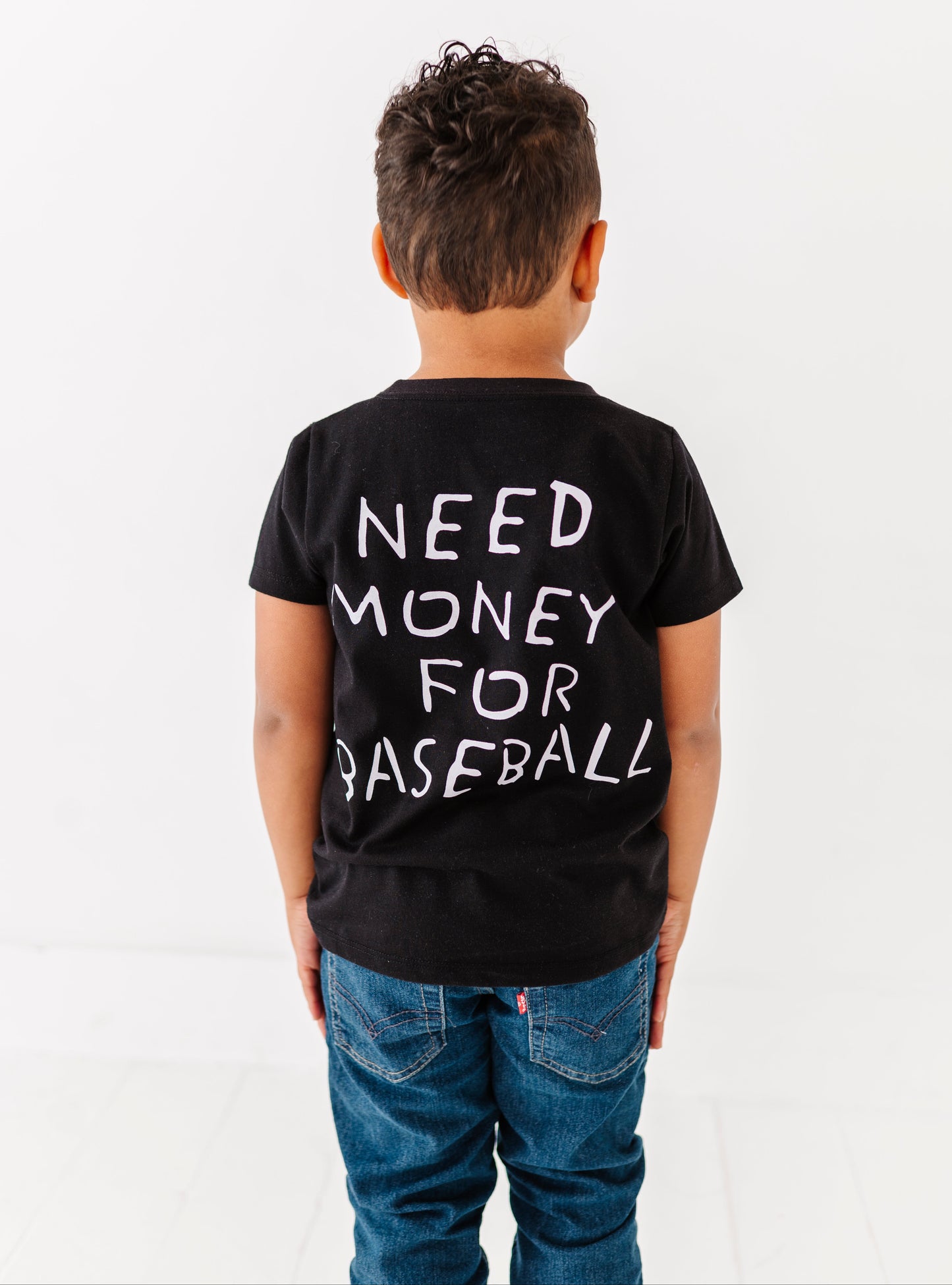 Child wearing a black t-shirt with 'NEED MONEY FOR BASEBALL' text on the back, standing against a white background.