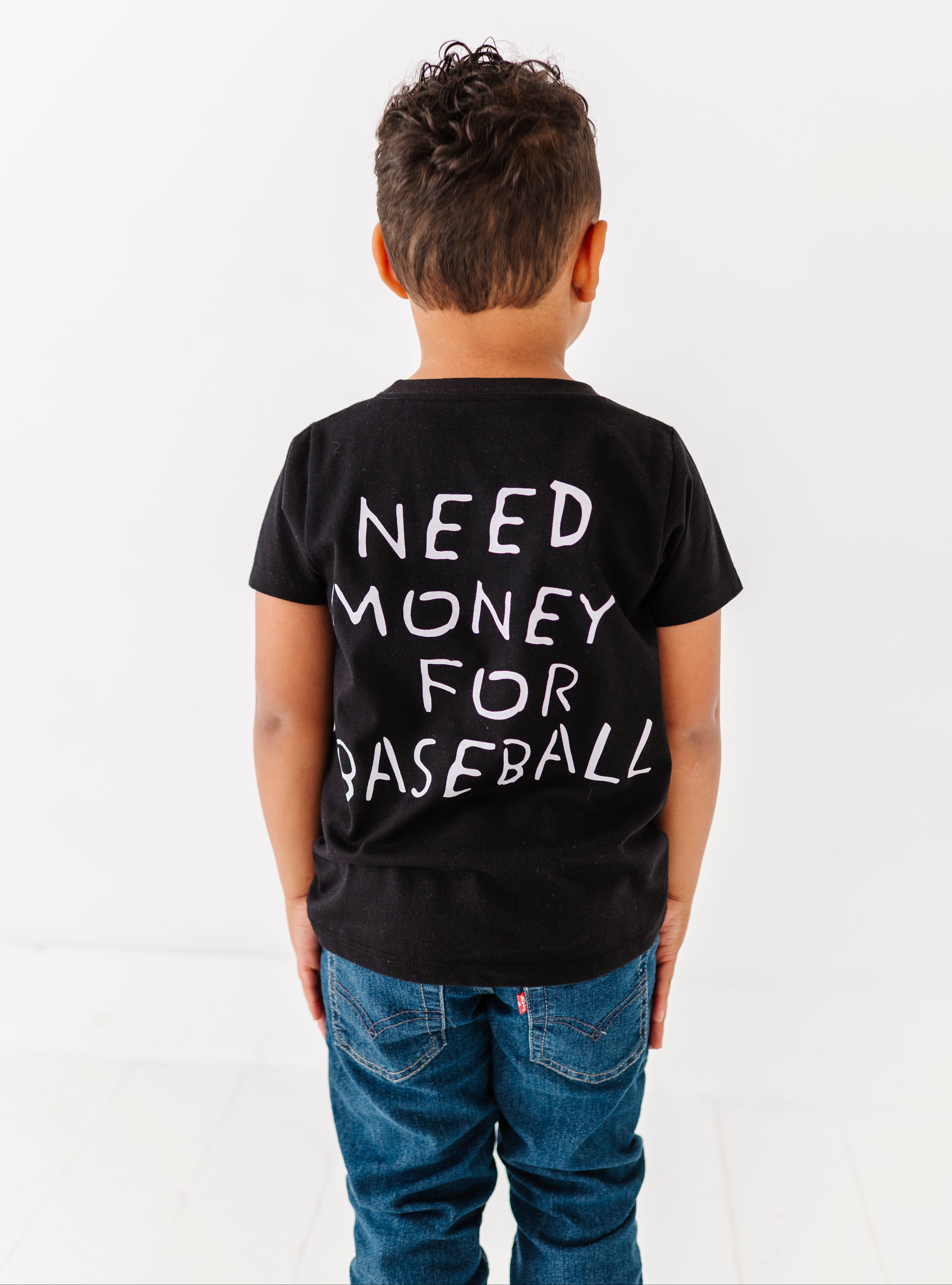 Child wearing a black t-shirt with 'NEED MONEY FOR BASEBALL' text on the back, standing against a white background.