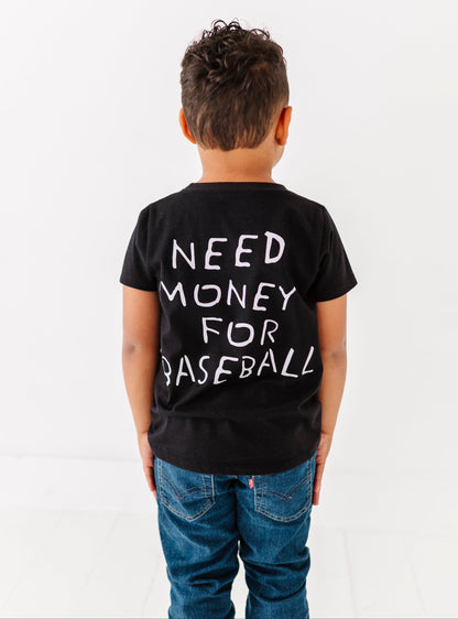 Child wearing a black t-shirt with 'NEED MONEY FOR BASEBALL' text on the back, standing against a white background.