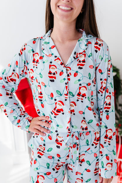 A woman wearing a light blue long-sleeve bamboo pajama set with a festive Christmas lights and Santa Claus print, holding a red gift bag.