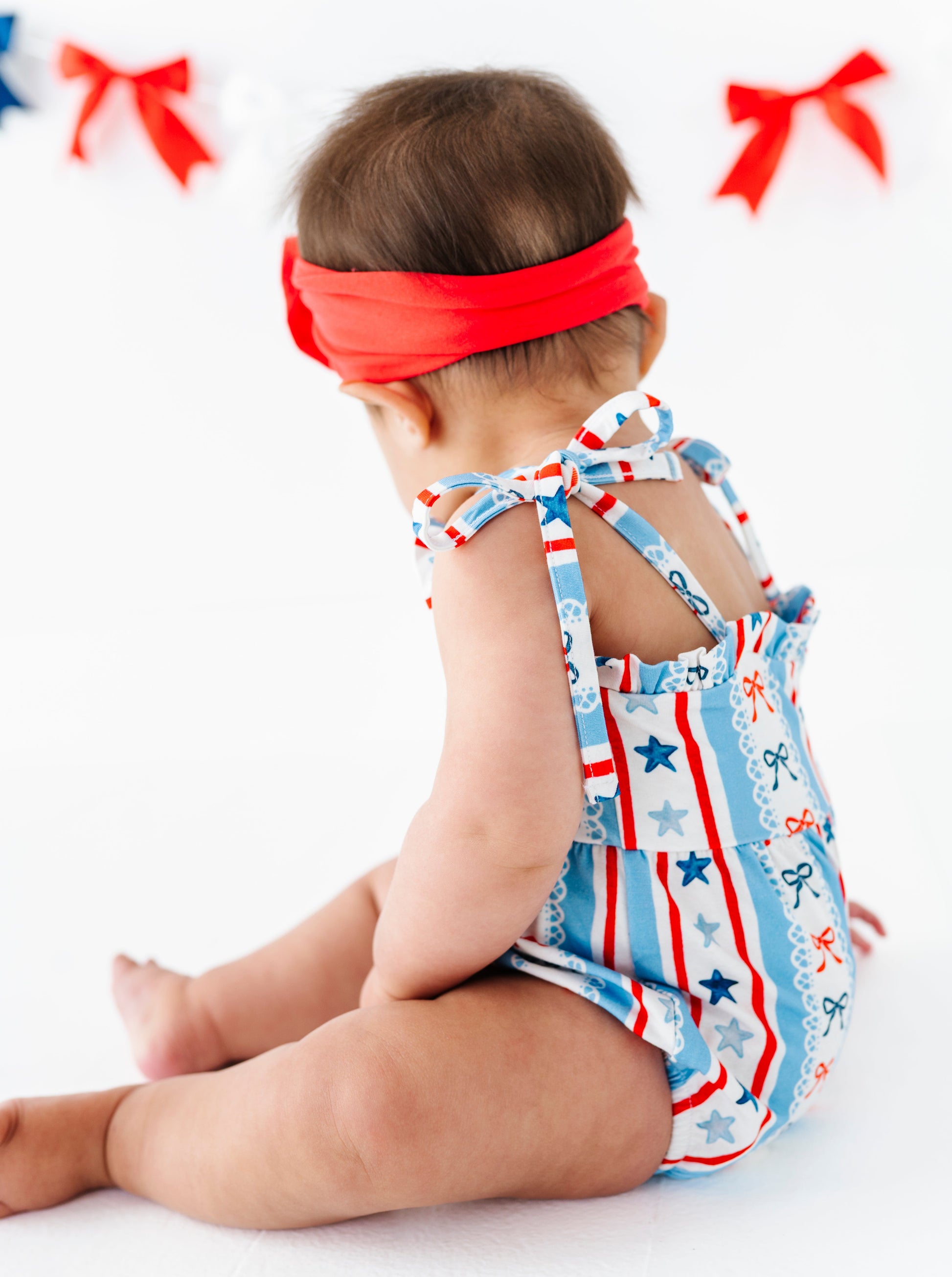 Baby wearing a red headband and a patriotic bamboo romper with star pattern on a white background