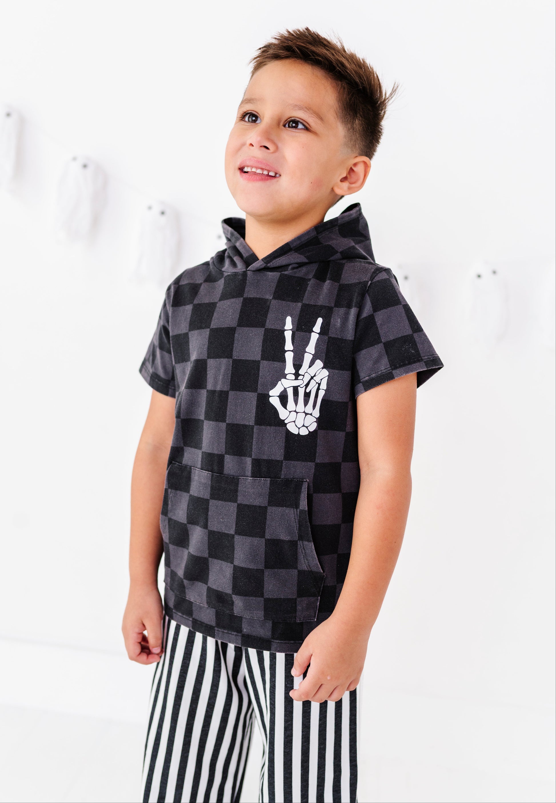 A young boy wearing a black and dark gray checkered short-sleeve hoodie with a white skeleton hand graphic on the chest, paired with black and white vertically striped pants, standing against a white wall.