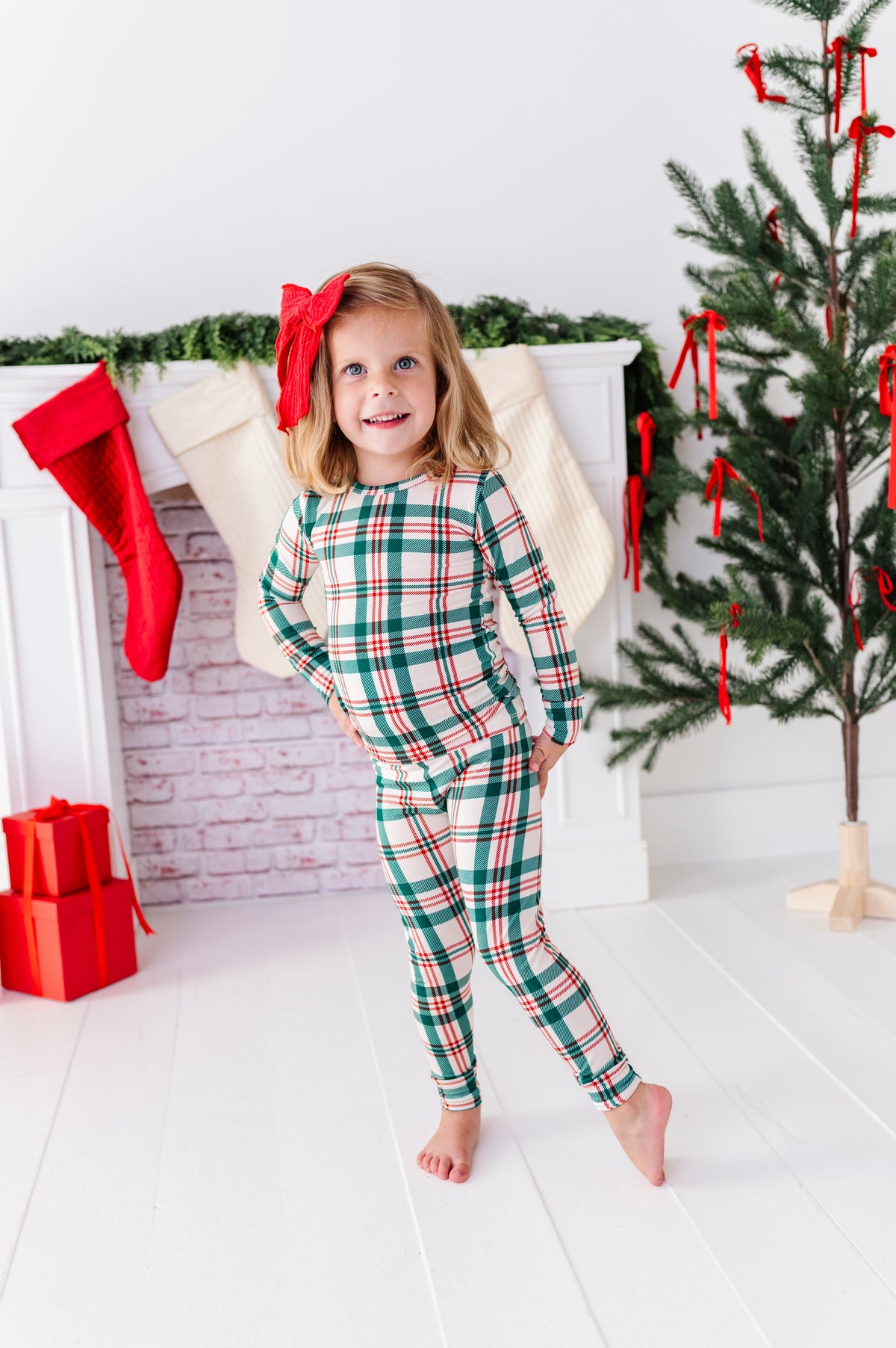 A little girl wearing a Classic Christmas plaid pajama set with a big red bow in her hair, standing in front of a decorated fireplace and a Christmas tree