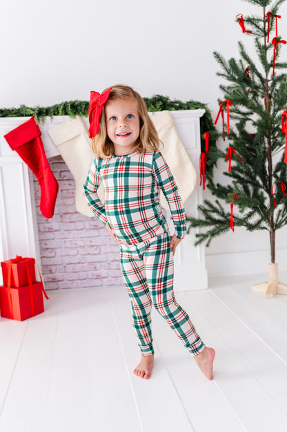 A little girl wearing a Classic Christmas plaid pajama set with a big red bow in her hair, standing in front of a decorated fireplace and a Christmas tree