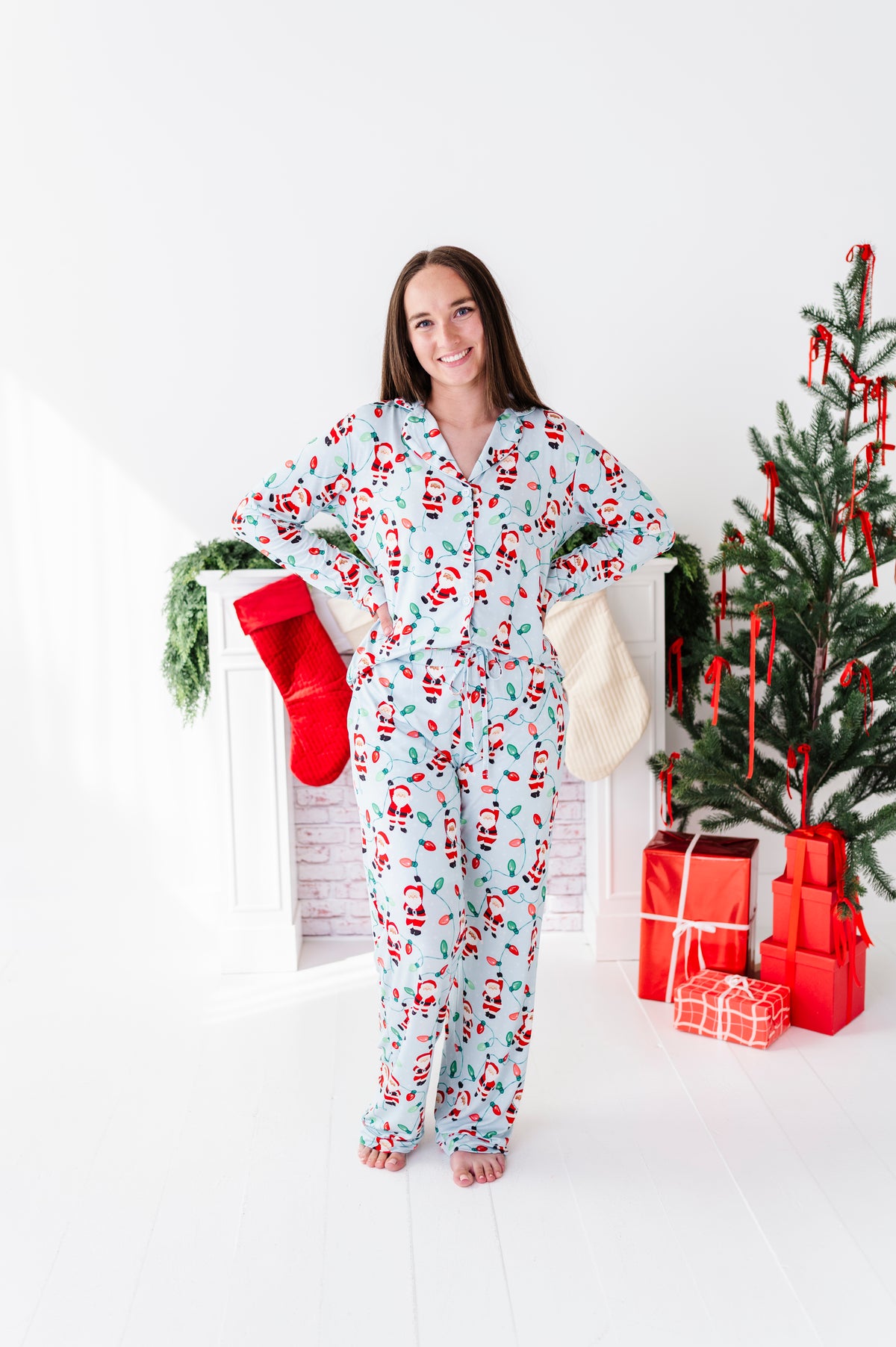 A woman wearing a light blue Christmas-themed bamboo pajama set with a Santa Claus print stands in a bright white room decorated for Christmas, holding a red stocking.