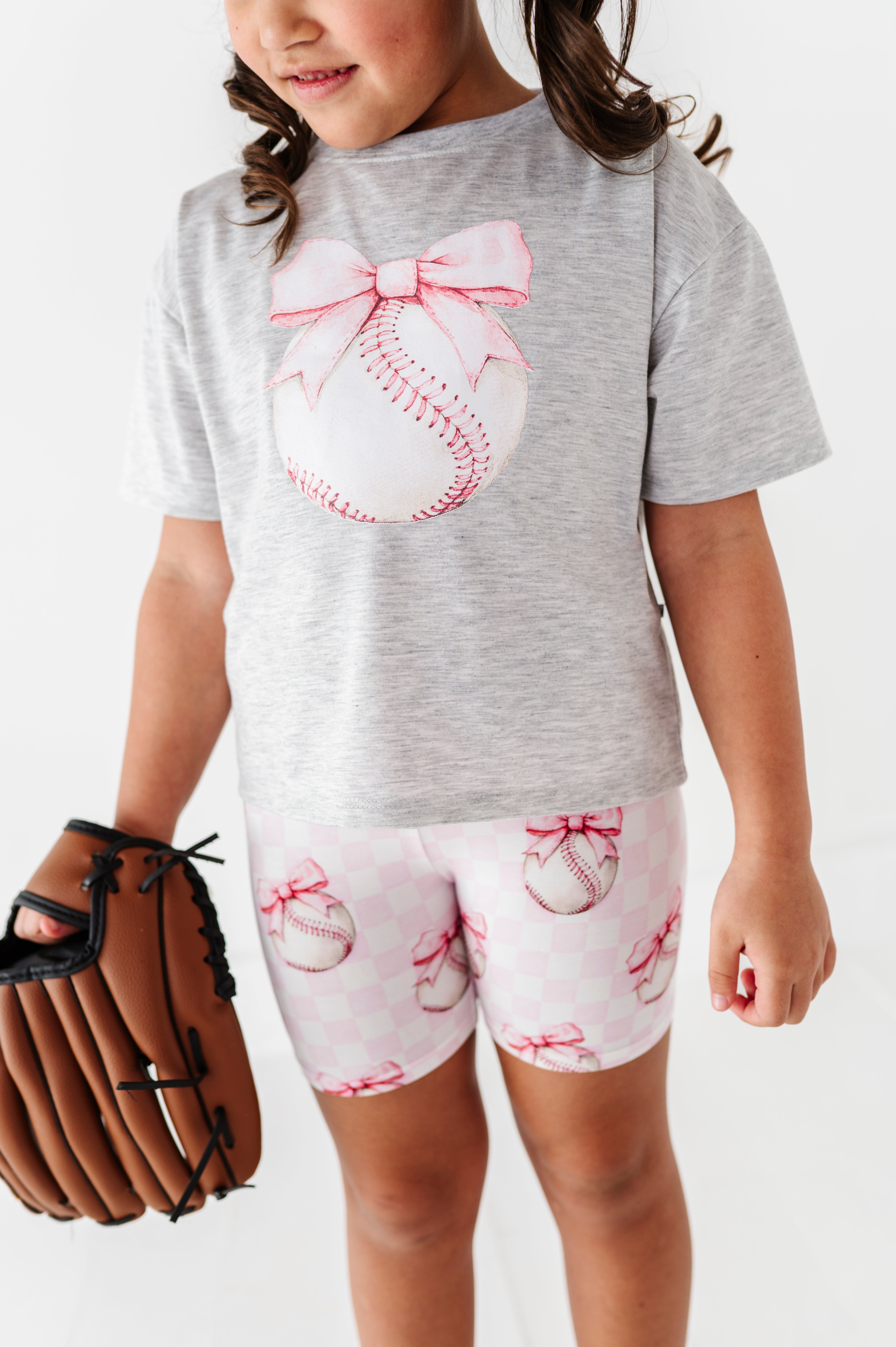 Young girl wearing a gray t-shirt with a baseball and bow design, holding a brown baseball glove.