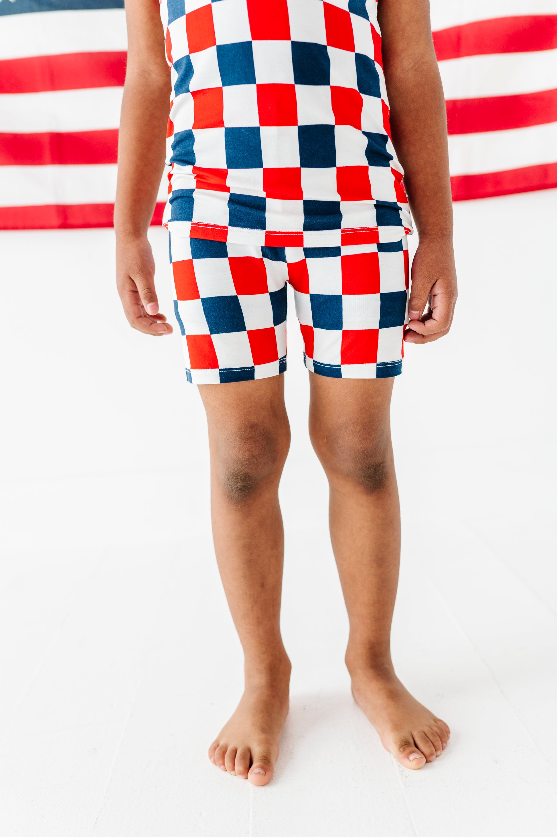 Little boy wearing red, white, and blue checkered bamboo pajama short set with an American flag in the background