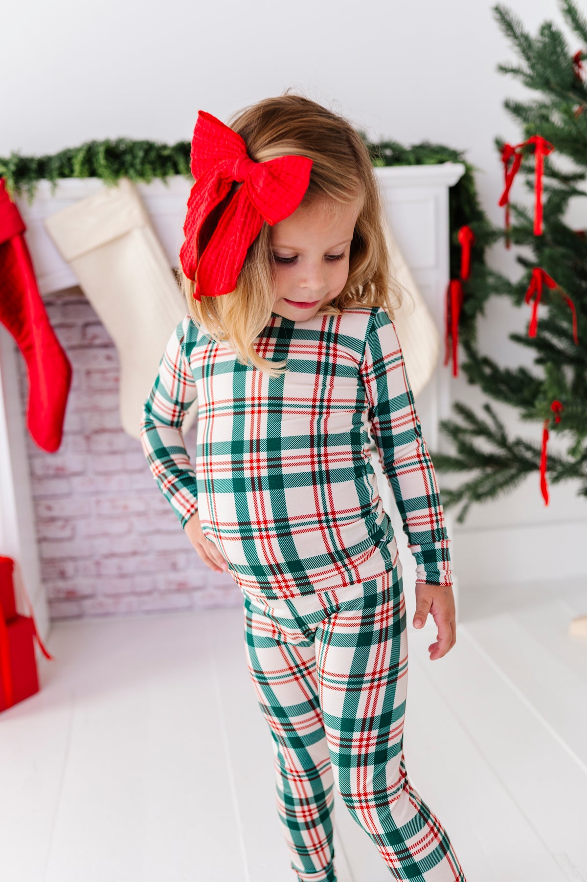 A little girl wearing a Classic Christmas plaid pajama set with a big red bow in her hair, standing in front of a decorated fireplace and a Christmas tree