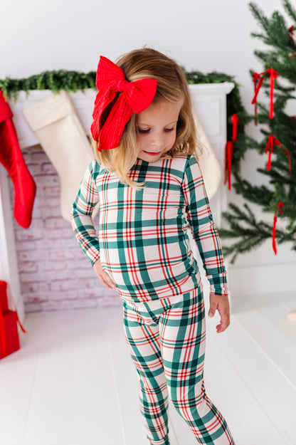 A little girl wearing a Classic Christmas plaid pajama set with a big red bow in her hair, standing in front of a decorated fireplace and a Christmas tree