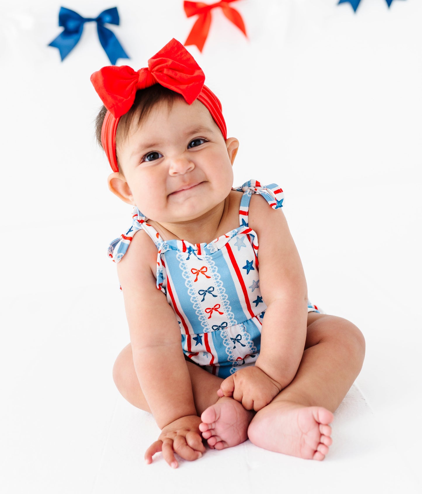 Baby wearing a red, white, and blue 4th of July-themed bamboo bubble romper with bows and a headband on a white background