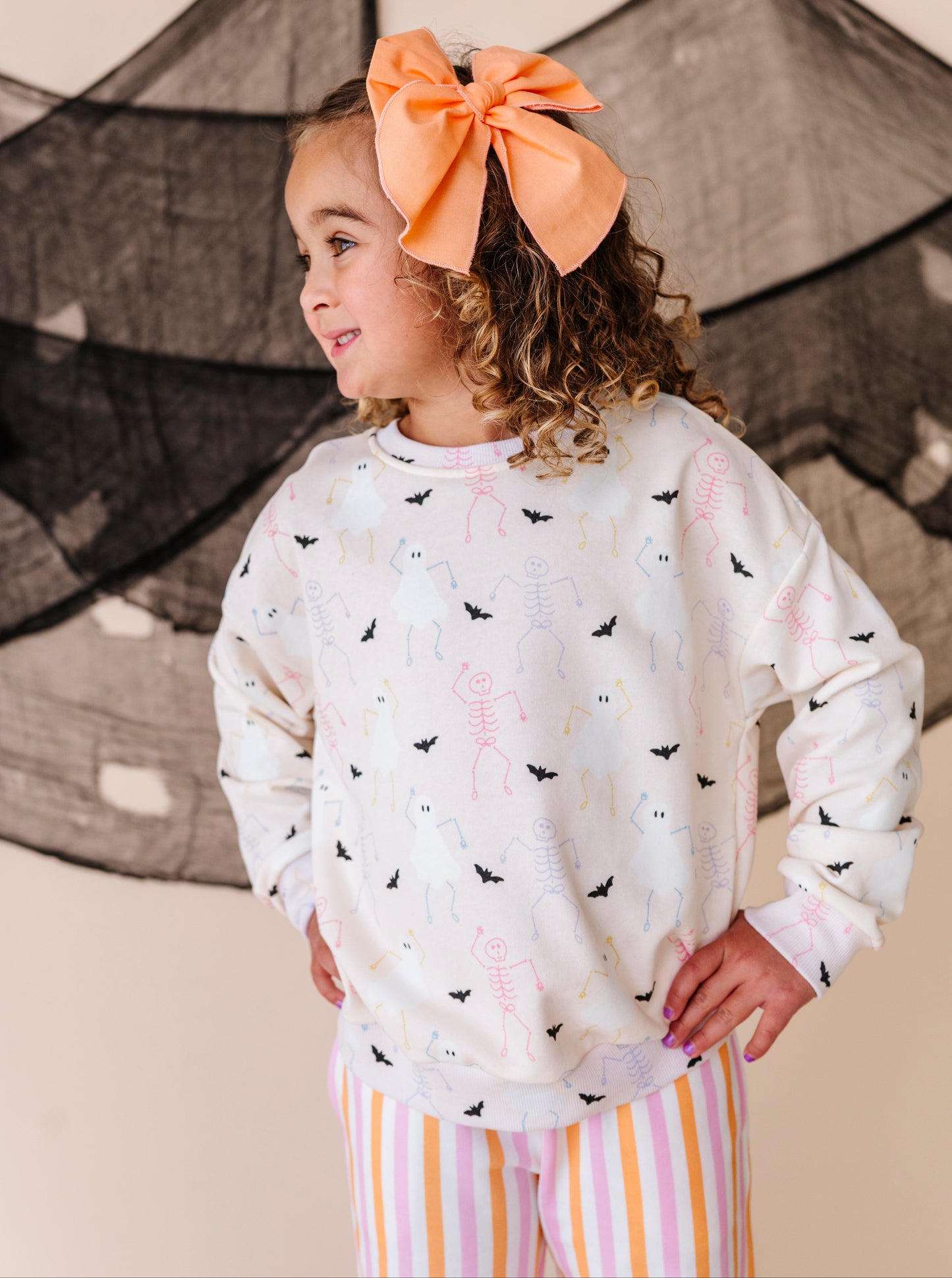 Young girl wearing a Halloween patterned sweatshirt  and striped pants with an orange bow in her hair, standing against a neutral background.