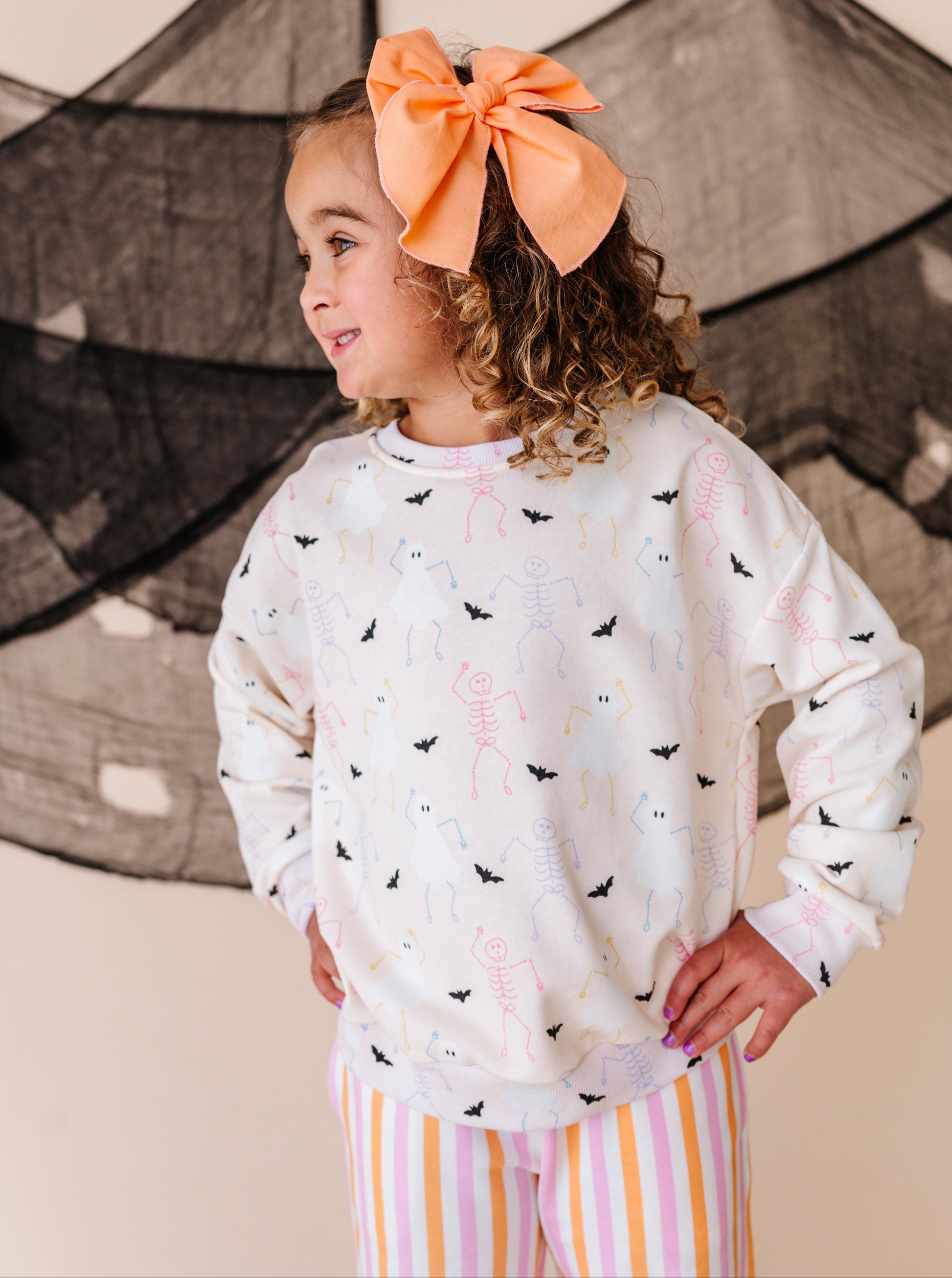 Young girl wearing a Halloween patterned sweatshirt  and striped pants with an orange bow in her hair, standing against a neutral background.