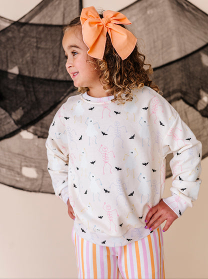 Young girl wearing a Halloween patterned sweatshirt  and striped pants with an orange bow in her hair, standing against a neutral background.