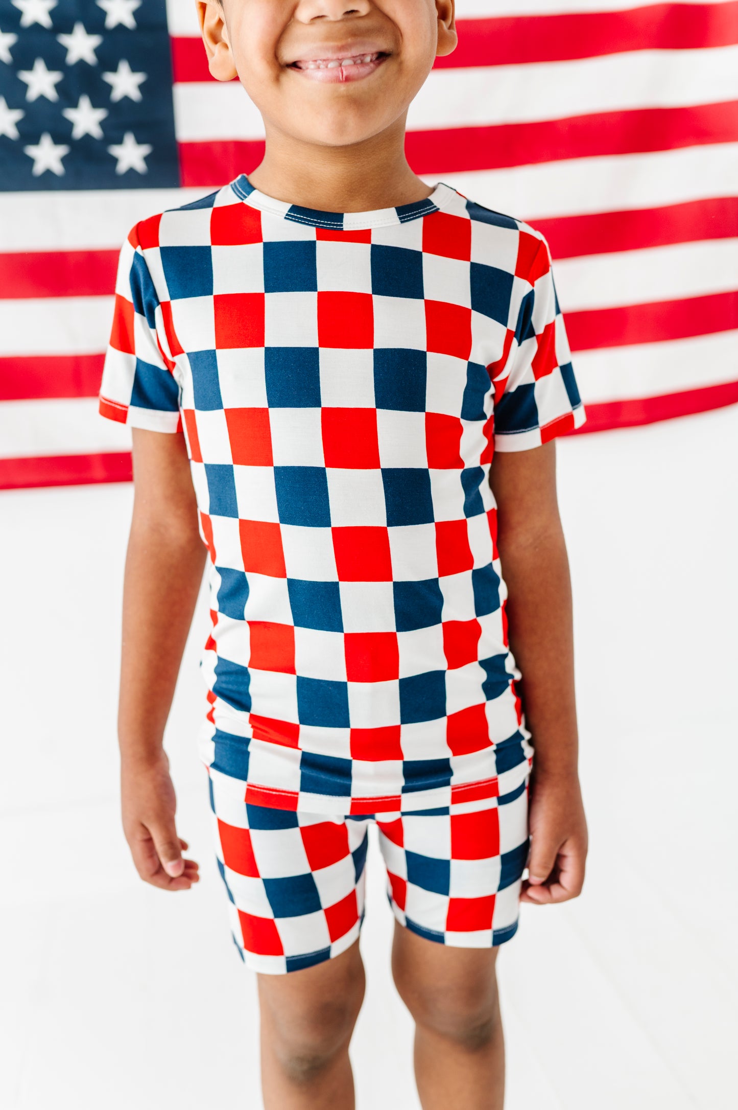 Child wearing a red, white, and blue checkered bamboo two piece pajama outfit in front of an American flag.