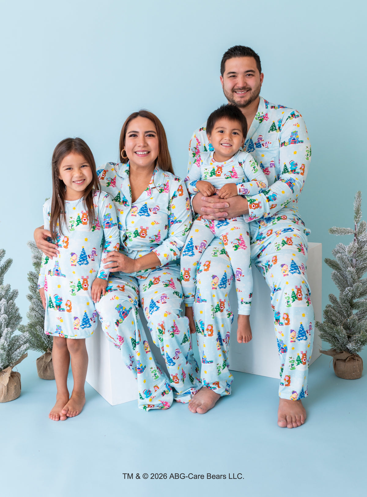 A family of four wearing matching light blue pajamas with a colorful Christmas tree print, posed together on a white block against a light blue background with small potted trees.