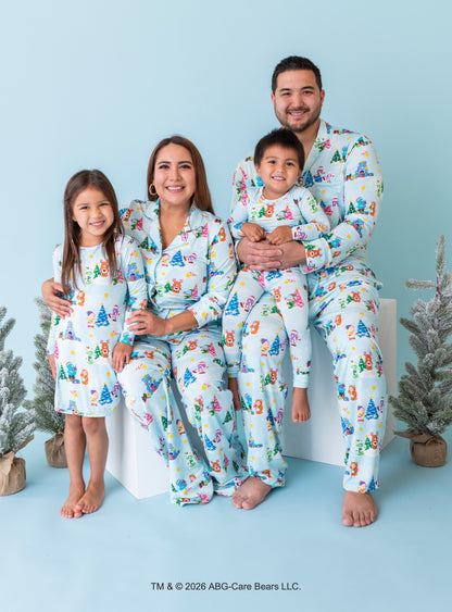 A family of four wearing matching light blue pajamas with a colorful Christmas tree print, posed together on a white block against a light blue background with small potted trees.