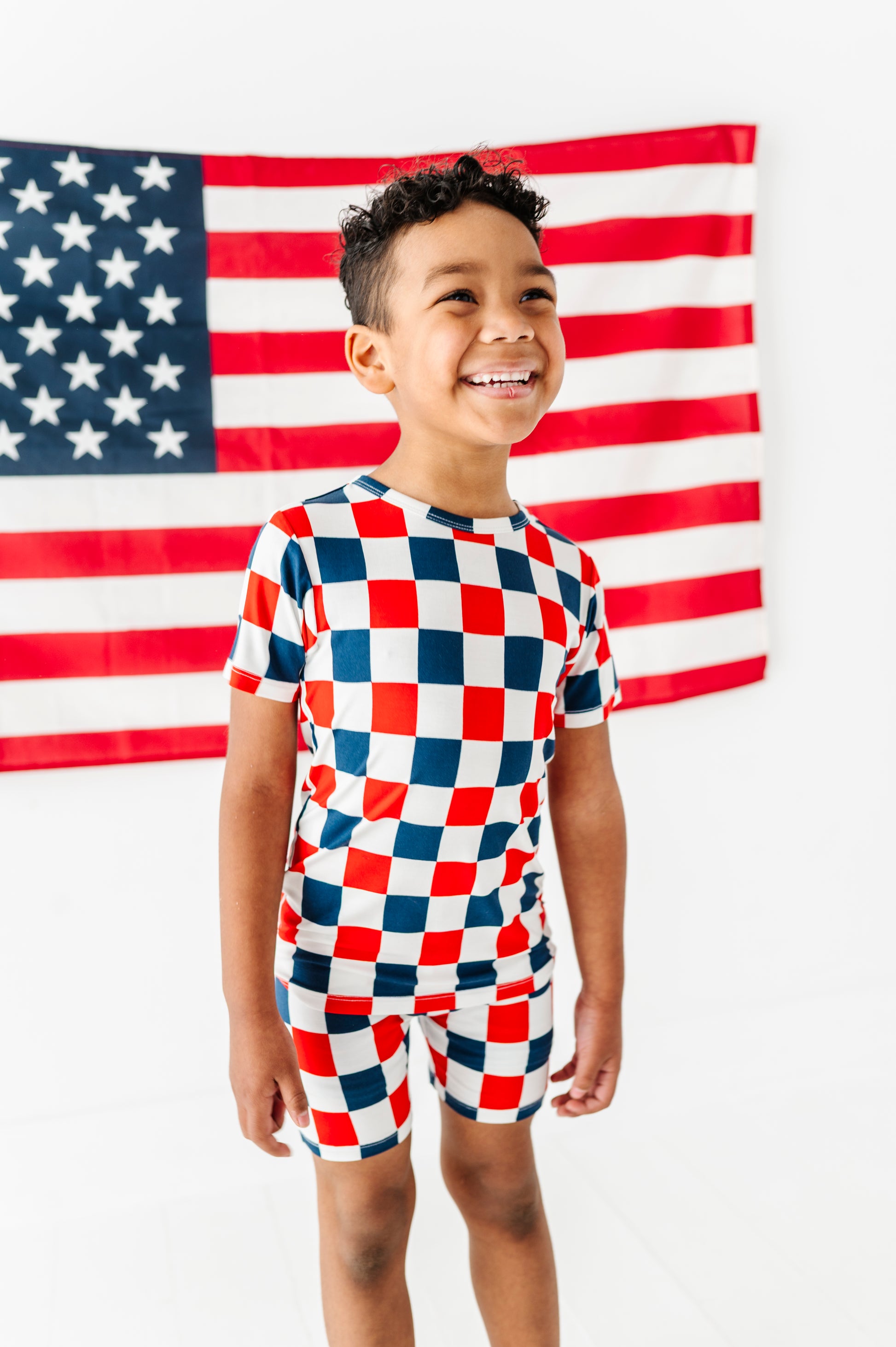 Child wearing a red, white, and blue checkered bamboo pajama outfit in front of an American flag.