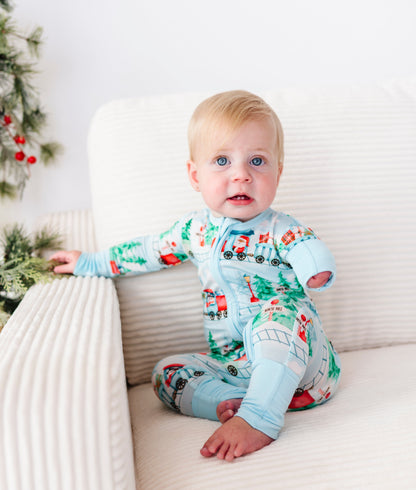 A baby sitting on a cream ribbed armchair wearing a light blue Christmas-themed bamboo sleeper with a train print, next to a decorated Christmas tree.