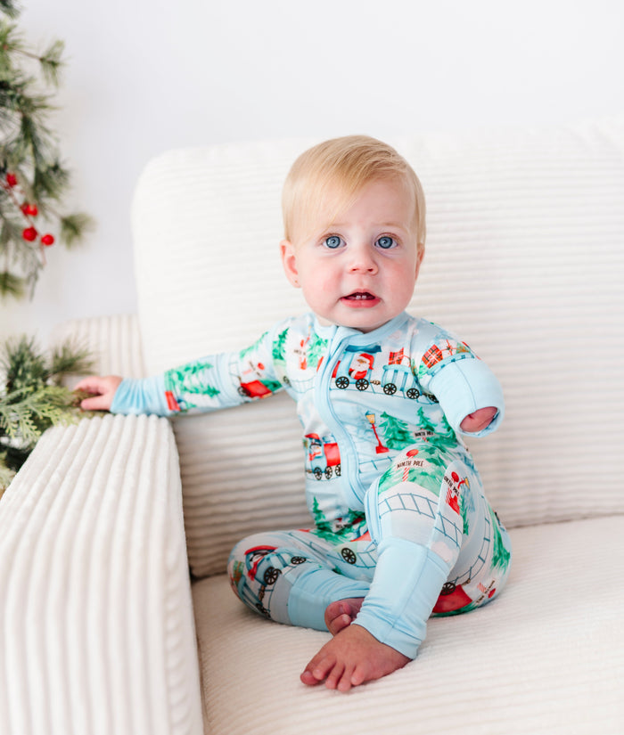 A baby sitting on a cream ribbed armchair wearing a light blue Christmas-themed bamboo sleeper with a train print, next to a decorated Christmas tree.