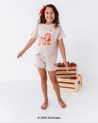 Young girl wearing a Strawberry Shortcake™ bamboo cotton beige outfit, standing next to a wooden crate filled with strawberries on a white background.