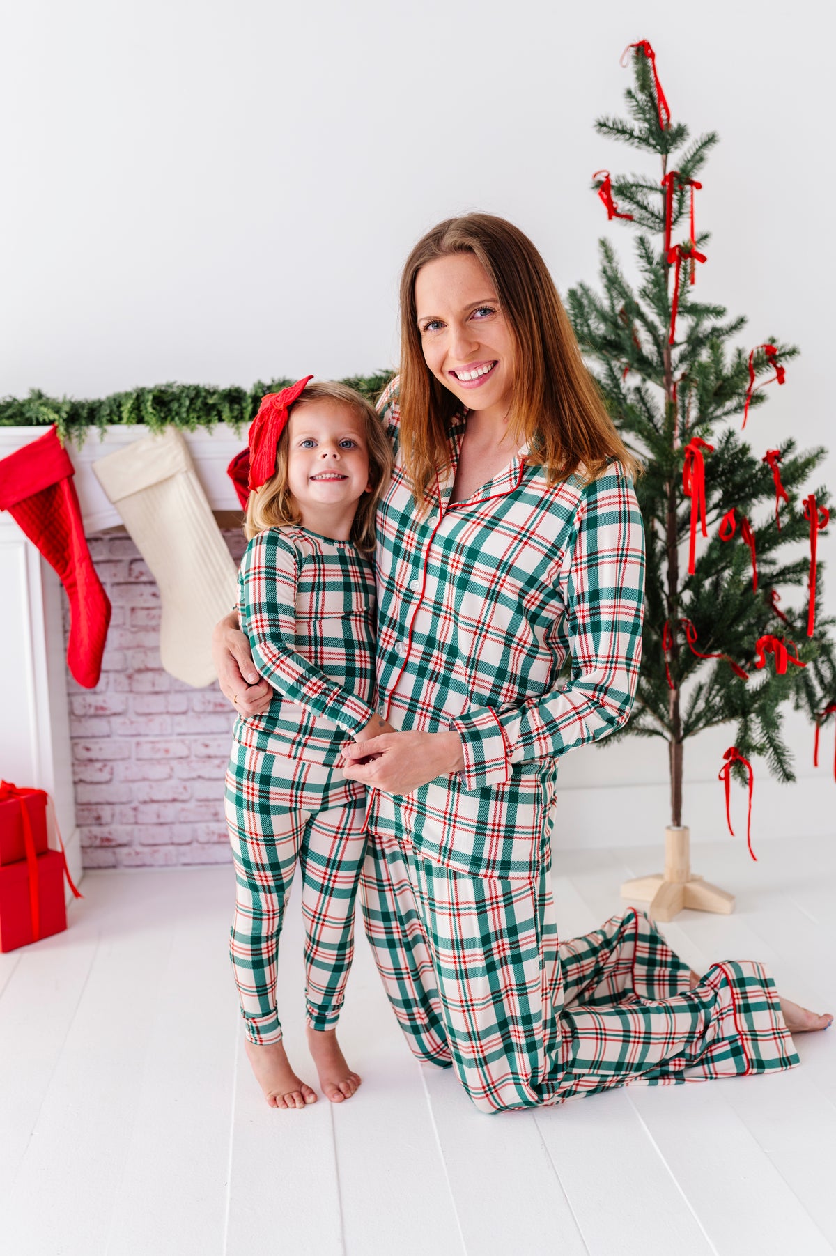 Mom and daughter standing in front of a Christmas-themed fireplace with stockings, wearing matching classic Christmas plaid pajamas