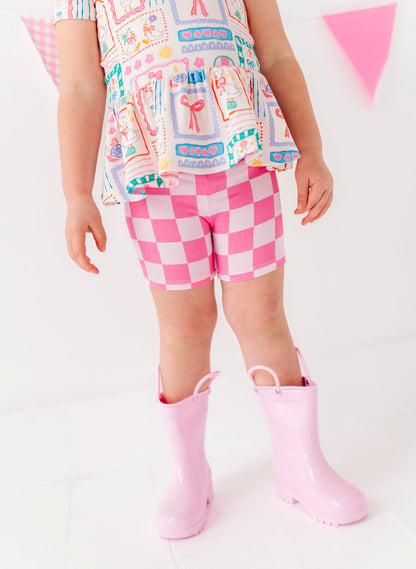 Child wearing a colorful top, pink checkered shorts, and pink rain boots on a white background
