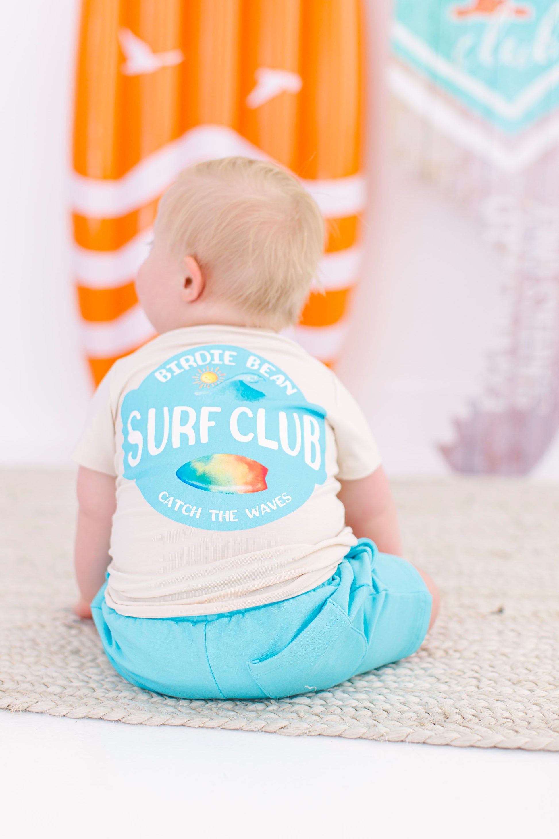 Baby sitting in front of a surfboard wearing a beige graphic tee with an ocean, beachy design