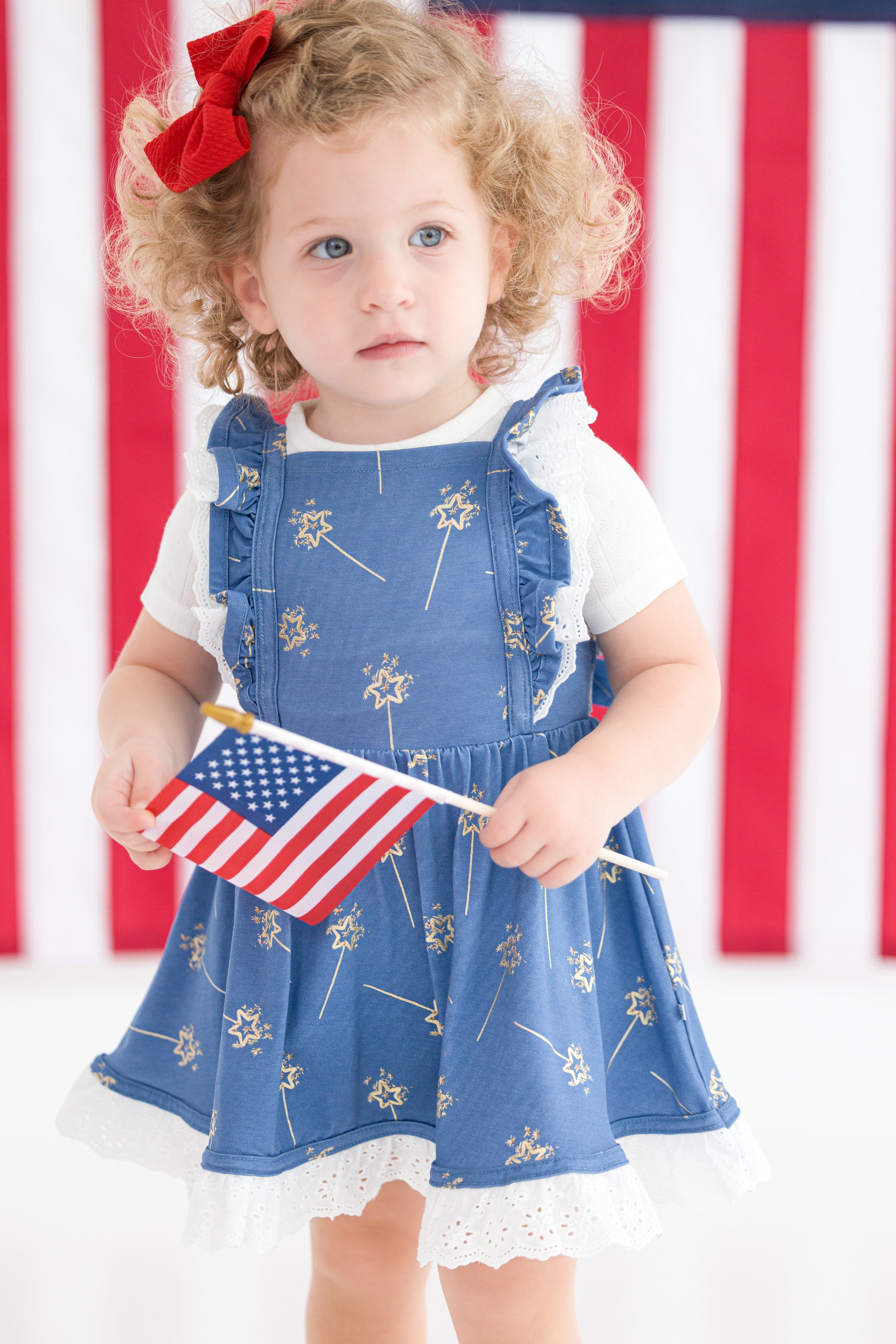Child wearing a blue dress with white sparklers and holding an American flag in front of an American flag background